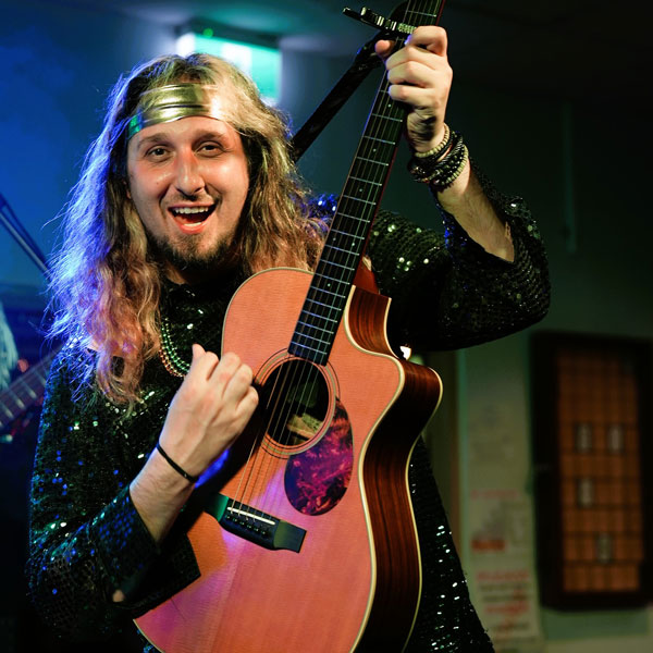 Smiling male musician with long hair and a headband playing an acoustic guitar on stage.