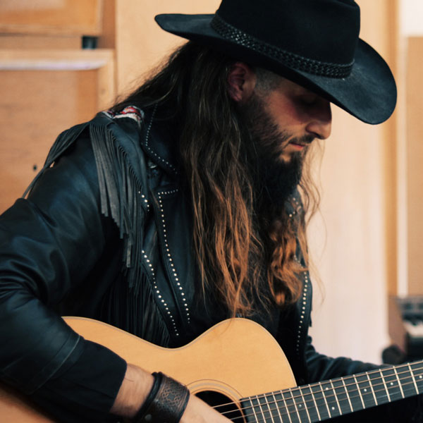 Man with long hair wearing a black cowboy hat and leather jacket playing an acoustic guitar indoors.