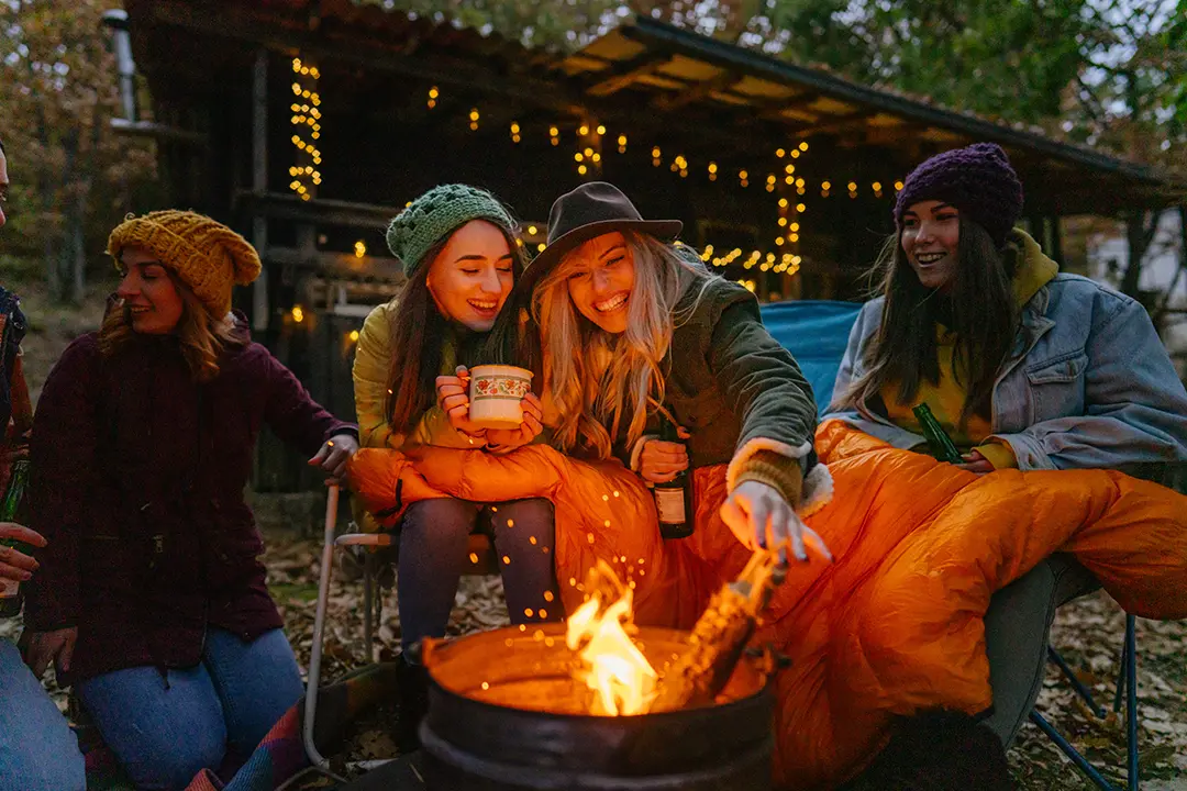 Group of friends sitting outdoors around a campfire in the evening, wearing warm clothing and smiling.