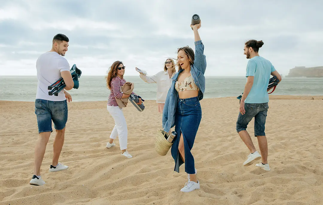 Five friends laughing and dancing on a sandy beach with a Bluetooth speaker near the ocean on a cloudy day.