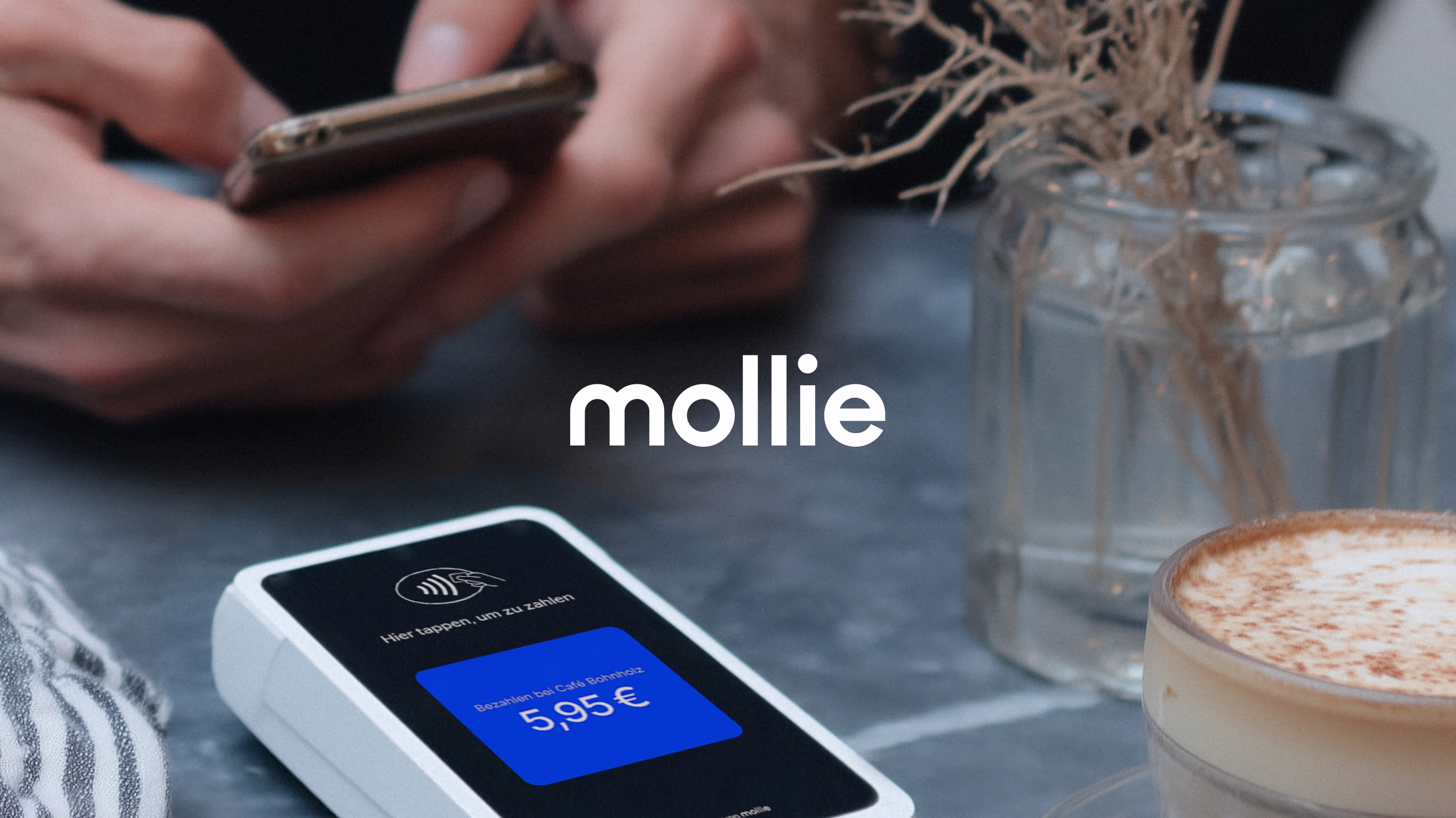 Nahaufnahme eines kontaktlosen Zahlungsterminals mit dem Preis 5,95 Euro in einem Café, Hände halten ein Smartphone im Hintergrund, auf dem Tisch eine Tasse Kaffee und ein Glas mit getrockneten Zweigen.