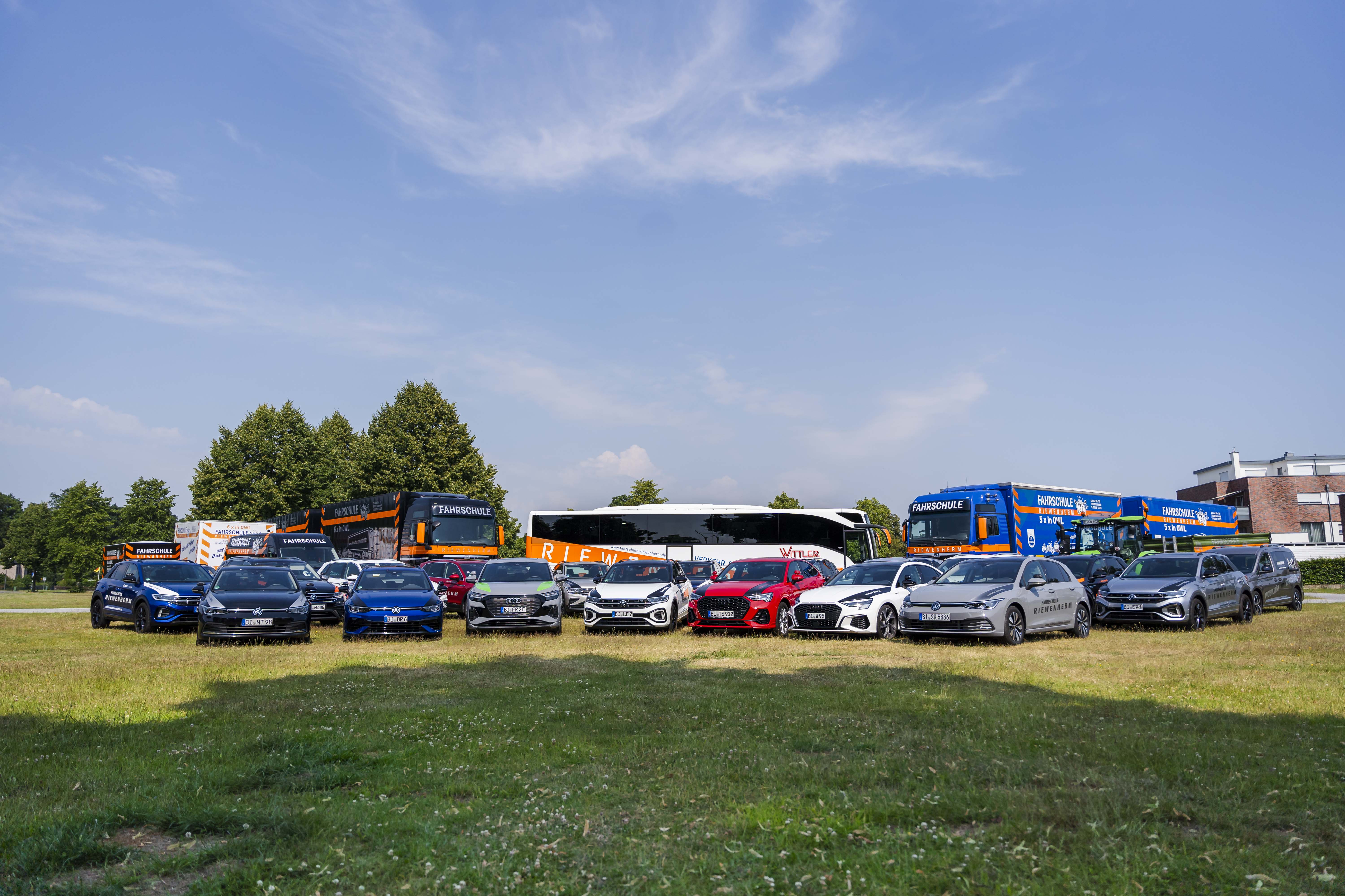 Verschiedene Fahrzeuge wie Autos, Busse und Lastwagen der Fahrschule Riewenherm auf einer grünen Wiese unter blauem Himmel.