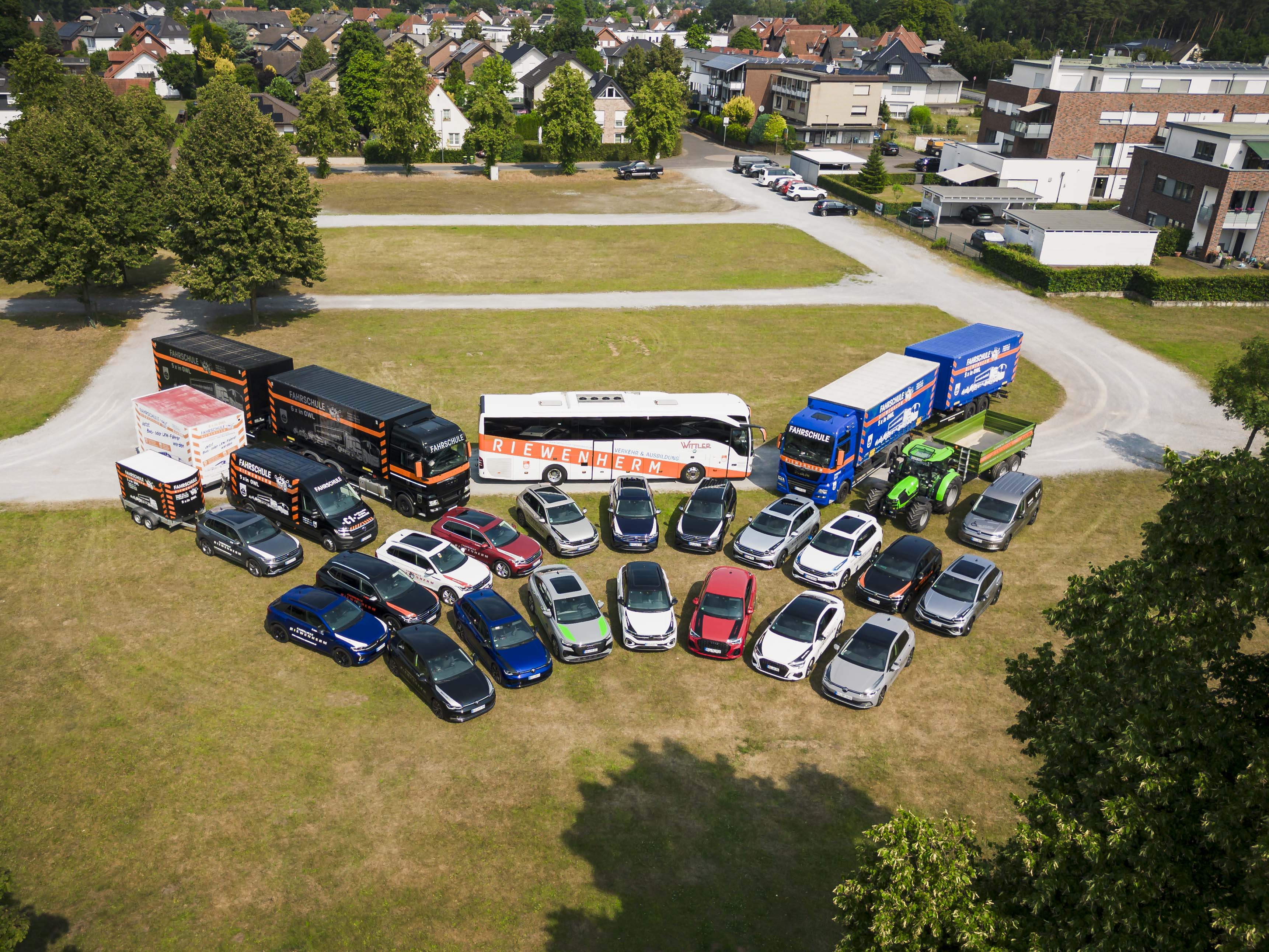 Flugaufnahme eines großen Fuhrparks mit verschiedenen Fahrzeugen, darunter LKW, Bus, Traktor und zahlreiche Autos auf einer Wiese in einer Wohngegend.