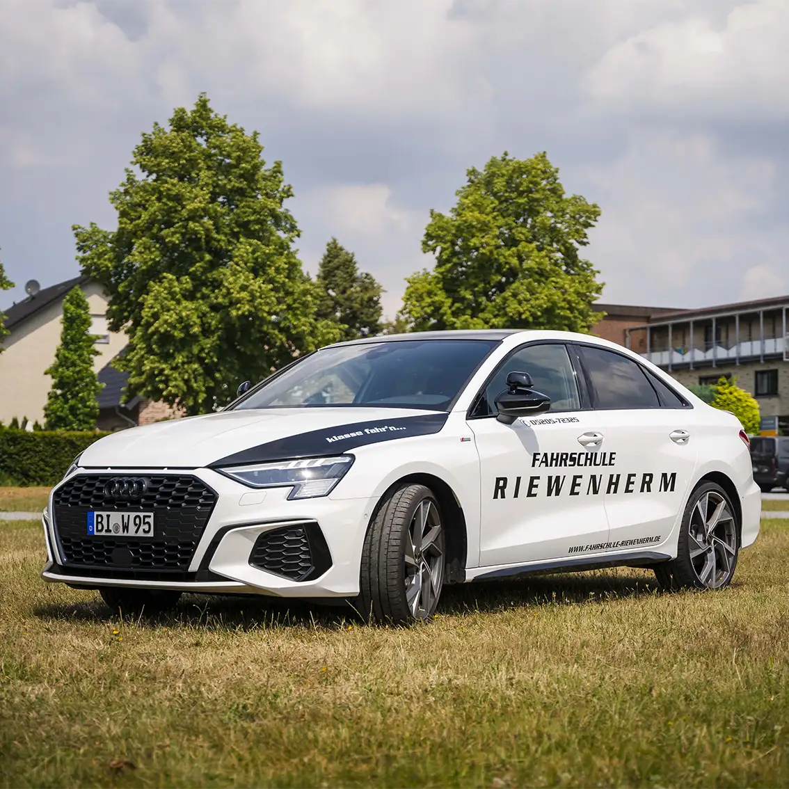 Weißes Audi-Fahrschulauto mit der Aufschrift ‚Fahrschule Riewenherm‘ auf der Seite, auf einer Wiese vor Bäumen und Häusern geparkt.