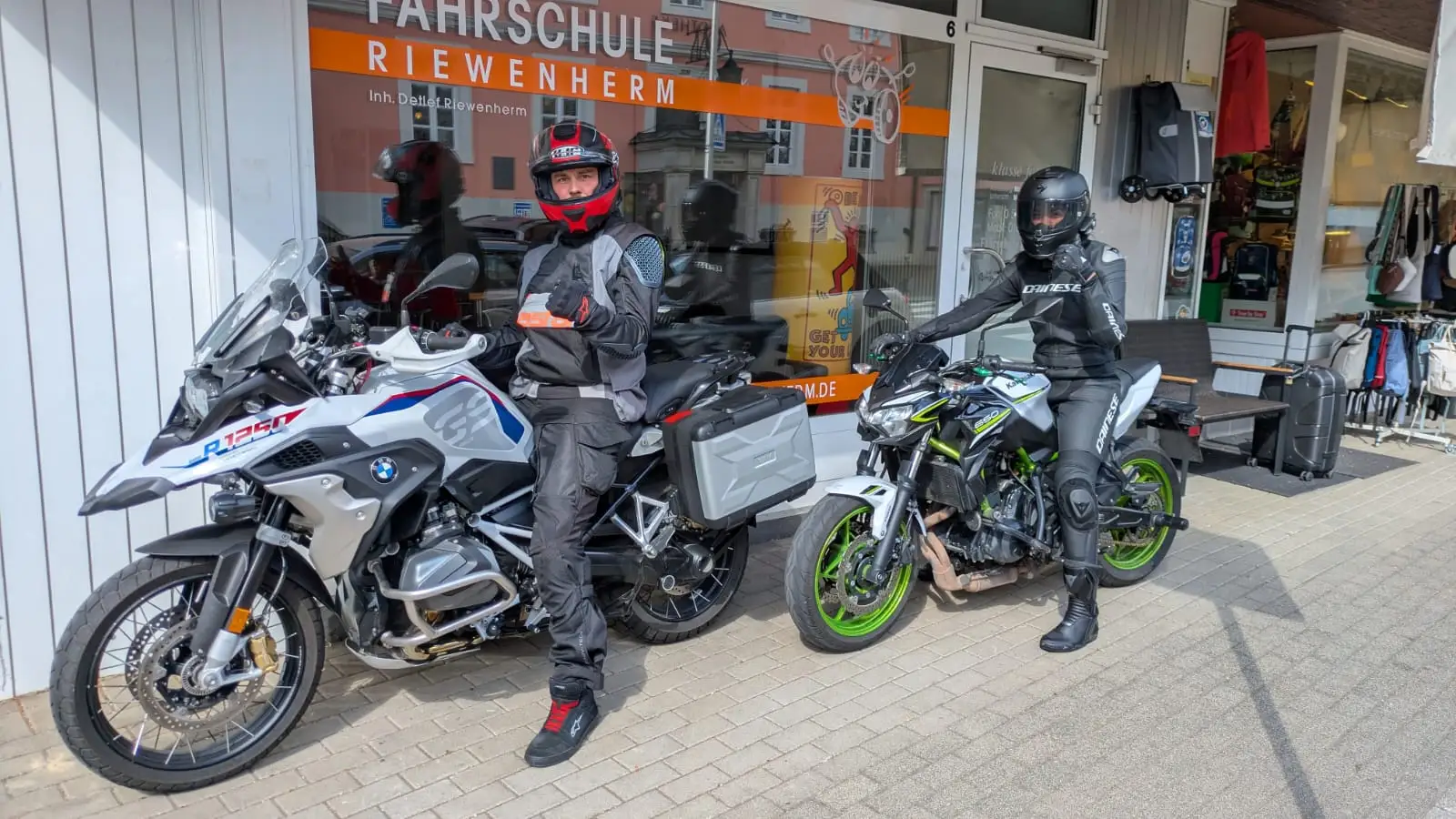 Zwei motorisierte Fahrer in Schutzkleidung posieren mit Motorrädern vor der Fahrschule Riewenherm.