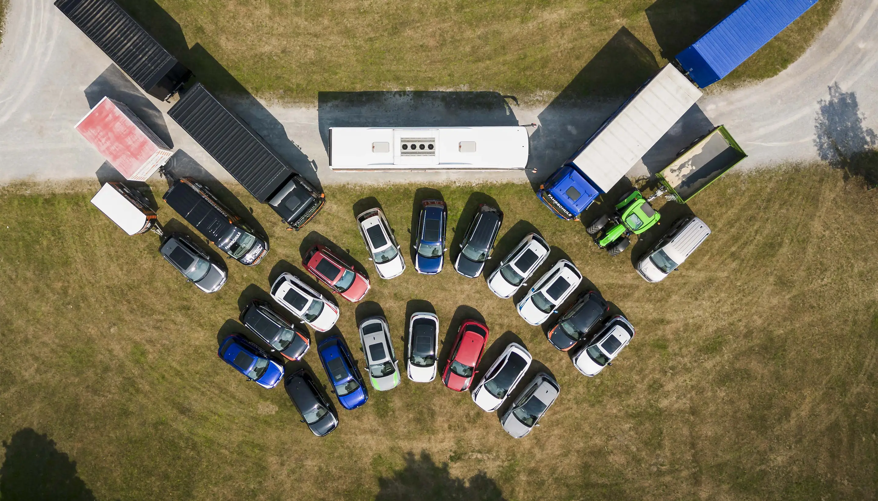 Vogelperspektive auf verschiedene Fahrzeuge, darunter Autos, LKWs, Bus und Traktor, auf einem Parkplatz mit Grasboden.