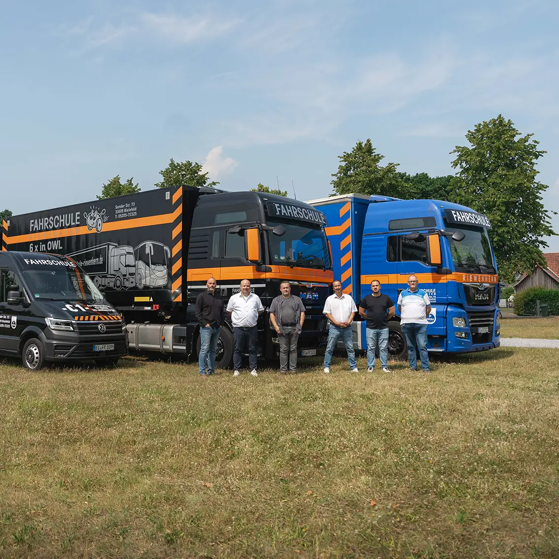 Sechs Männer stehen vor zwei Lastwagen und einem Van der Fahrschule Riewenherm auf einer Wiese unter blauem Himmel.