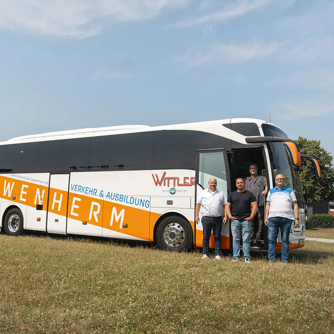 Vier Männer stehen vor einem weißen und orangefarbenen Bus der Fahrschule Riewenherm auf einer Wiese unter blauem Himmel.