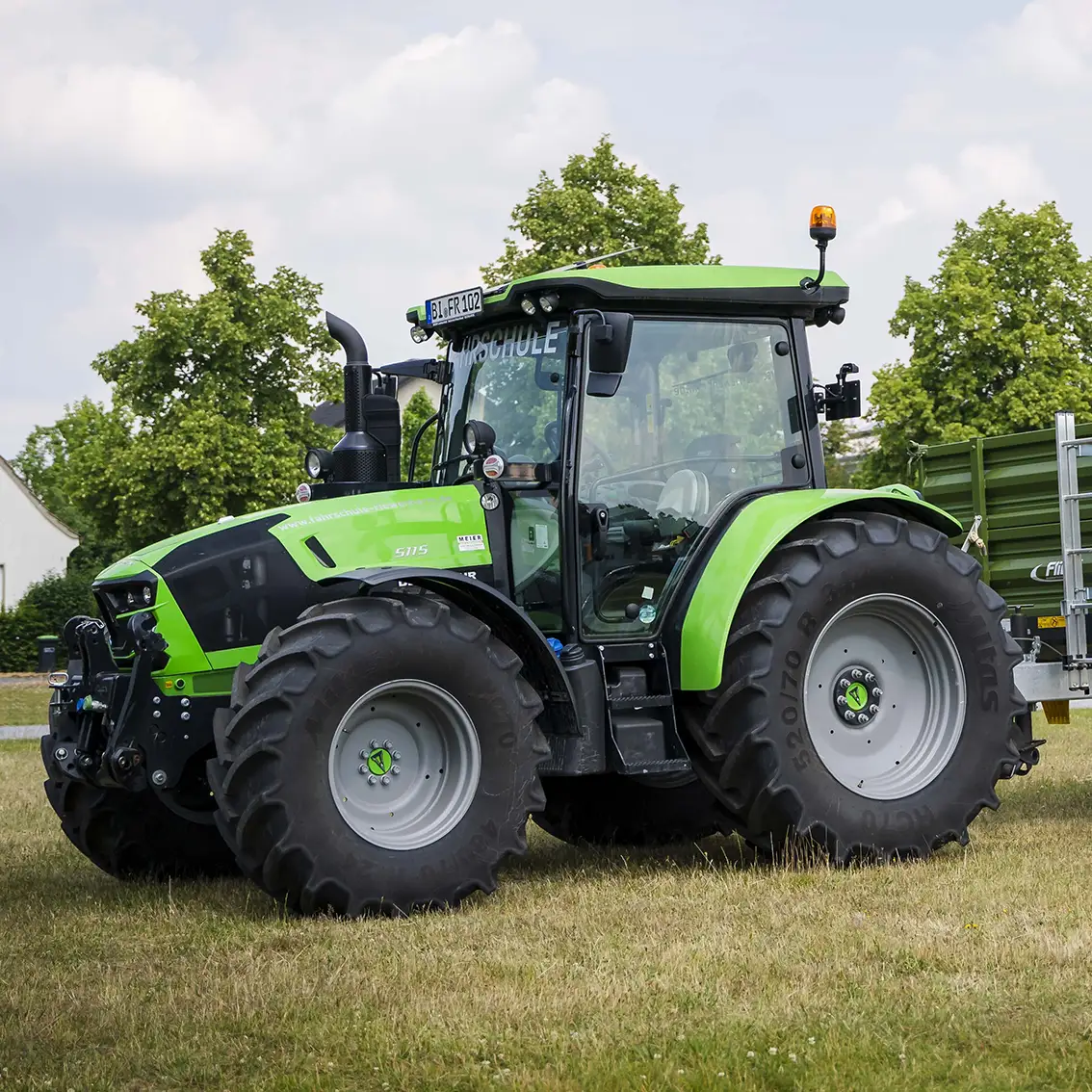 Grüner Traktor mit großem Reifen auf einer Wiese vor Bäumen und Häusern, seitlich aufgenommen.