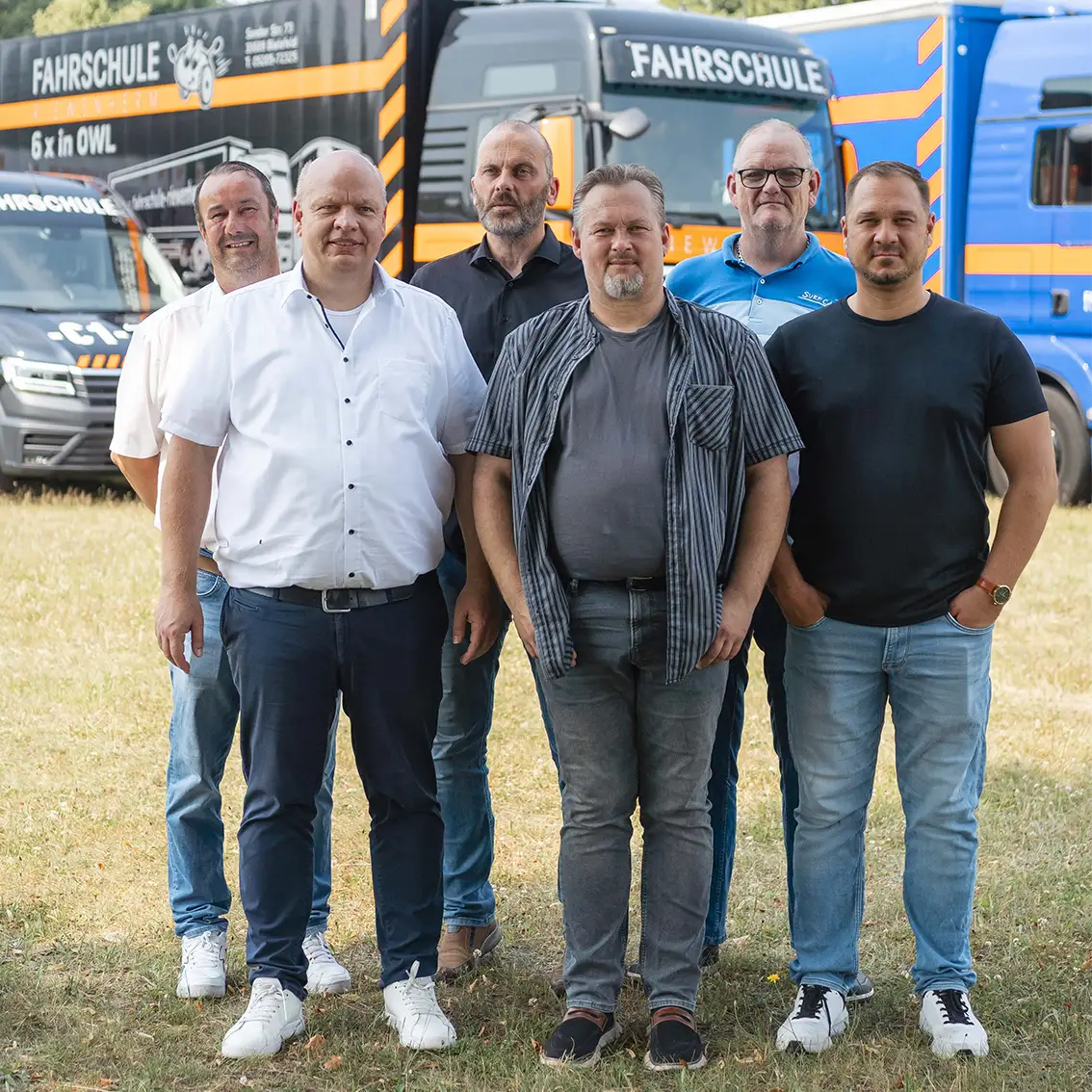 Sechs Männer stehen auf einer Wiese vor mehreren Fahrtschul-LKWs mit Aufschrift Fahrschule.