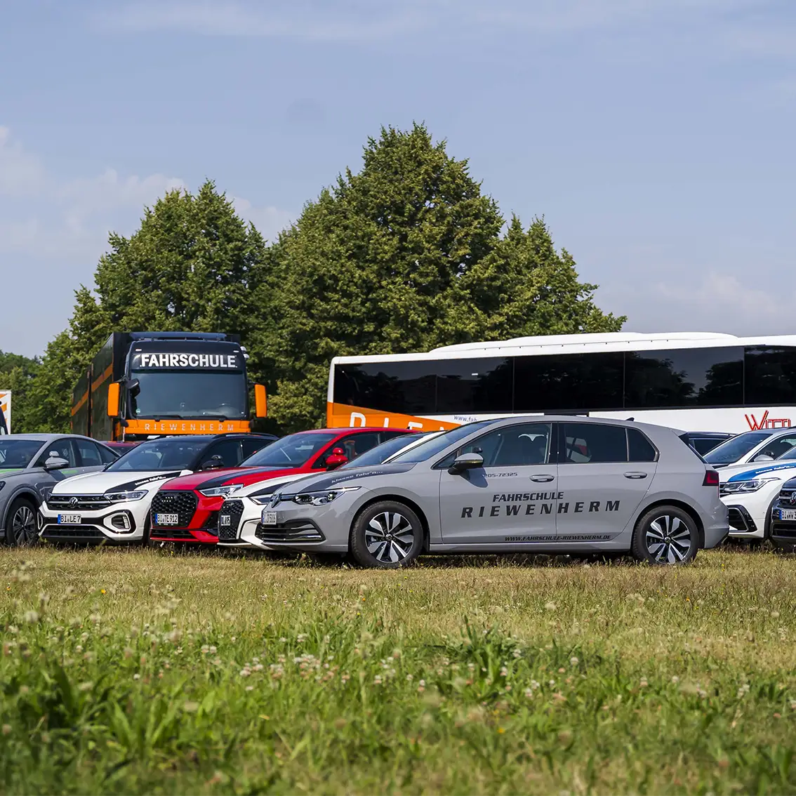 Mehrere Autos und zwei Busse mit dem Schriftzug Fahrschule Riewenherm auf einer Wiese vor Bäumen.