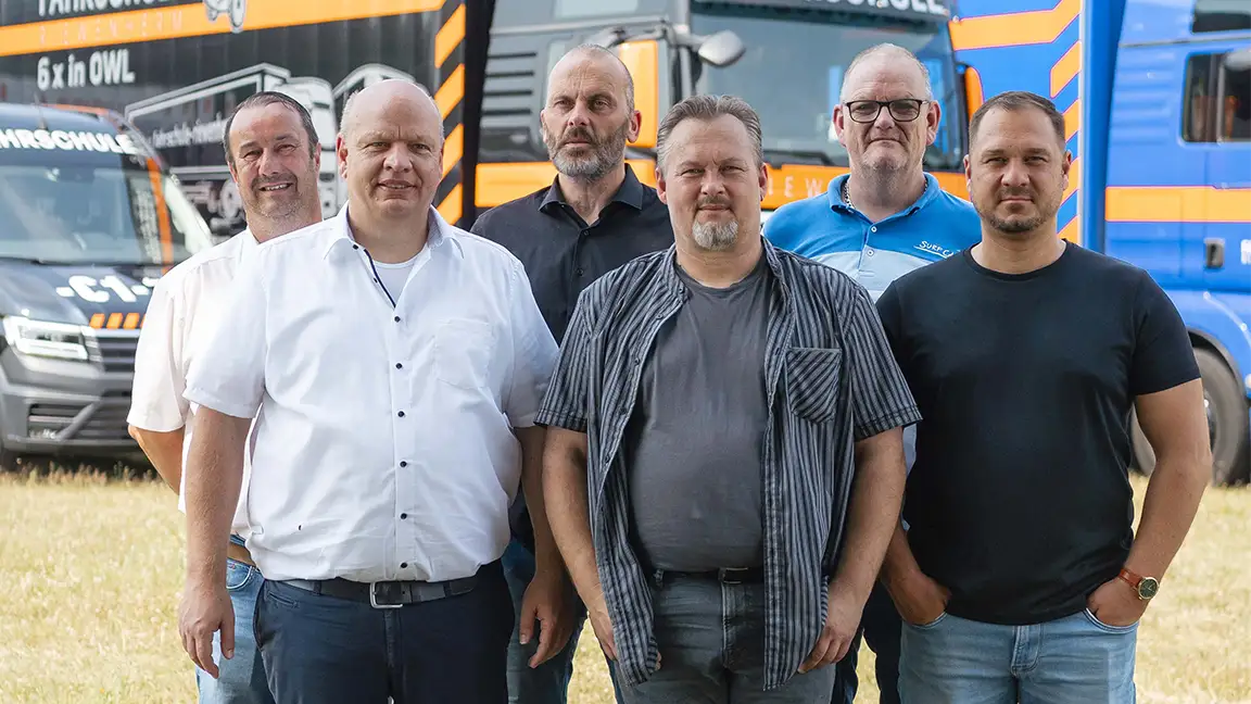 Sechs Männer stehen auf einem Feld vor Fahrzeugen mit Fahrschulwerbung.