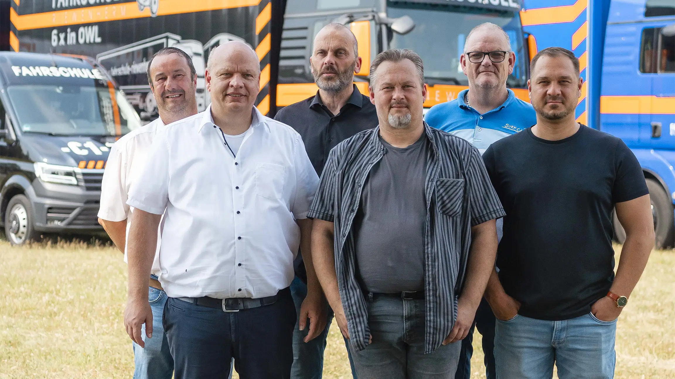 Sechs Männer stehen nebeneinander im Freien vor mehreren Lastwagen und Transportfahrzeugen.