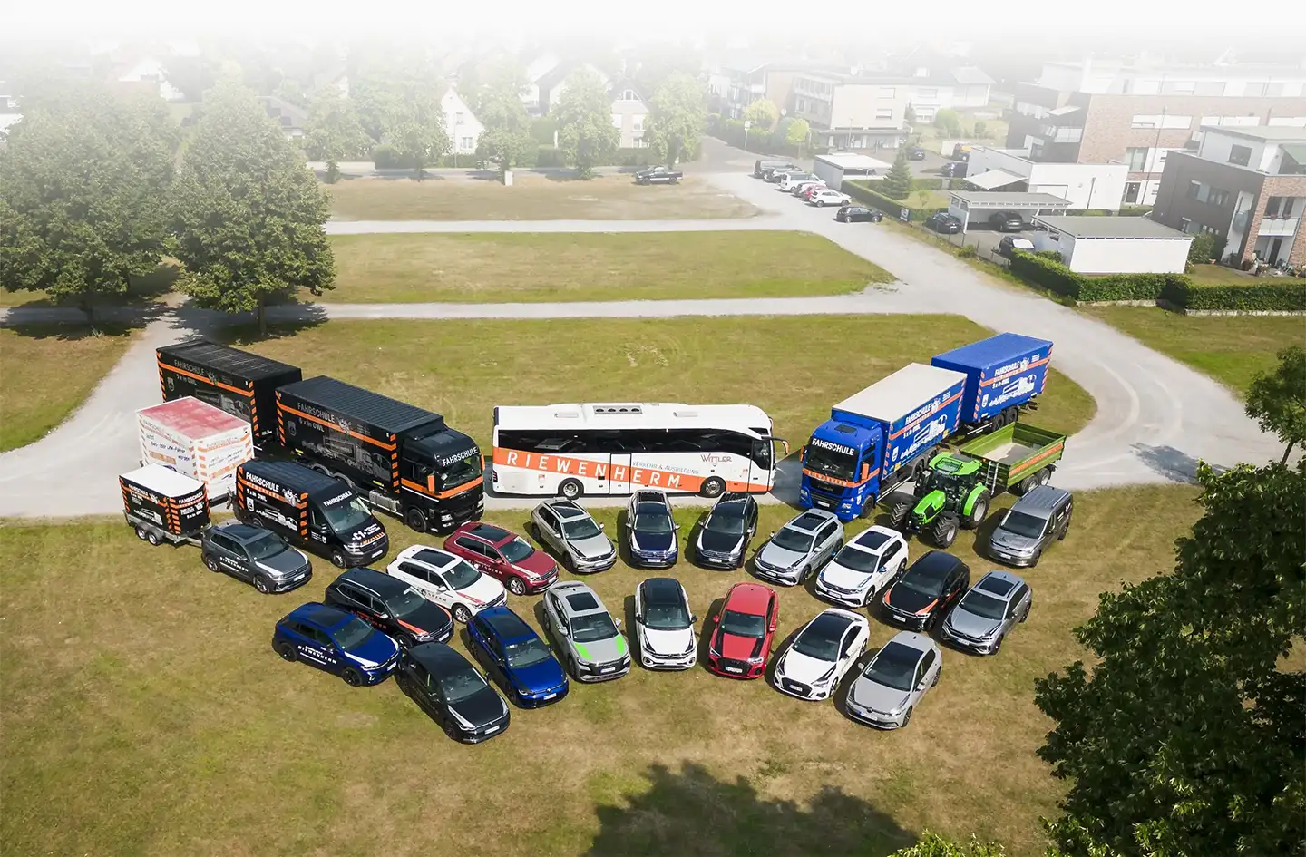 Verschiedene Fahrschulfahrzeuge, darunter Autos, Busse, Lastwagen und ein Traktor, auf einer Wiese in einer Wohngegend geparkt.