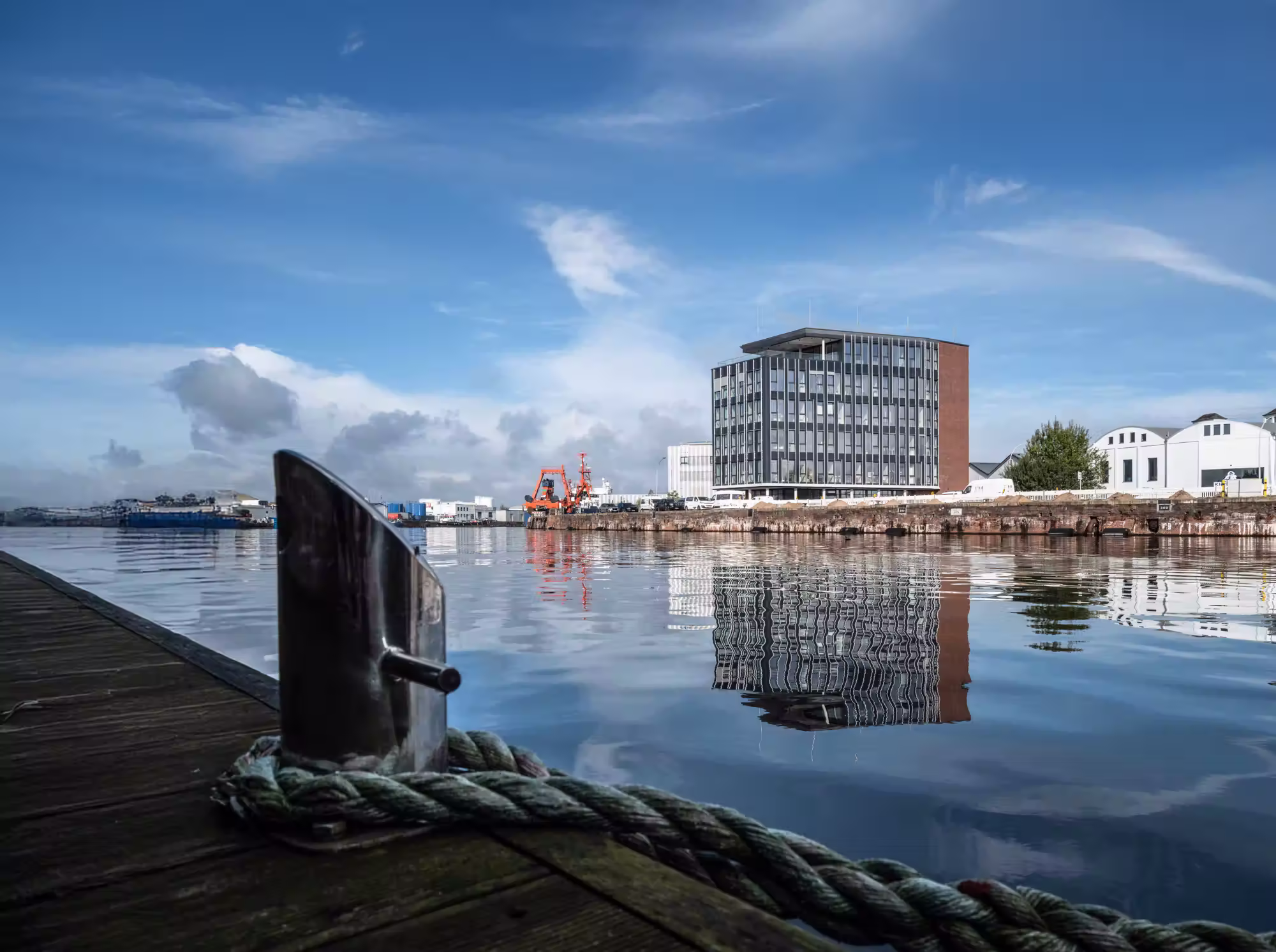 Blick vom hölzernen Hafensteg auf einen ruhigen Hafen mit spiegelglattem Wasser und modernen Gebäuden im Hintergrund.