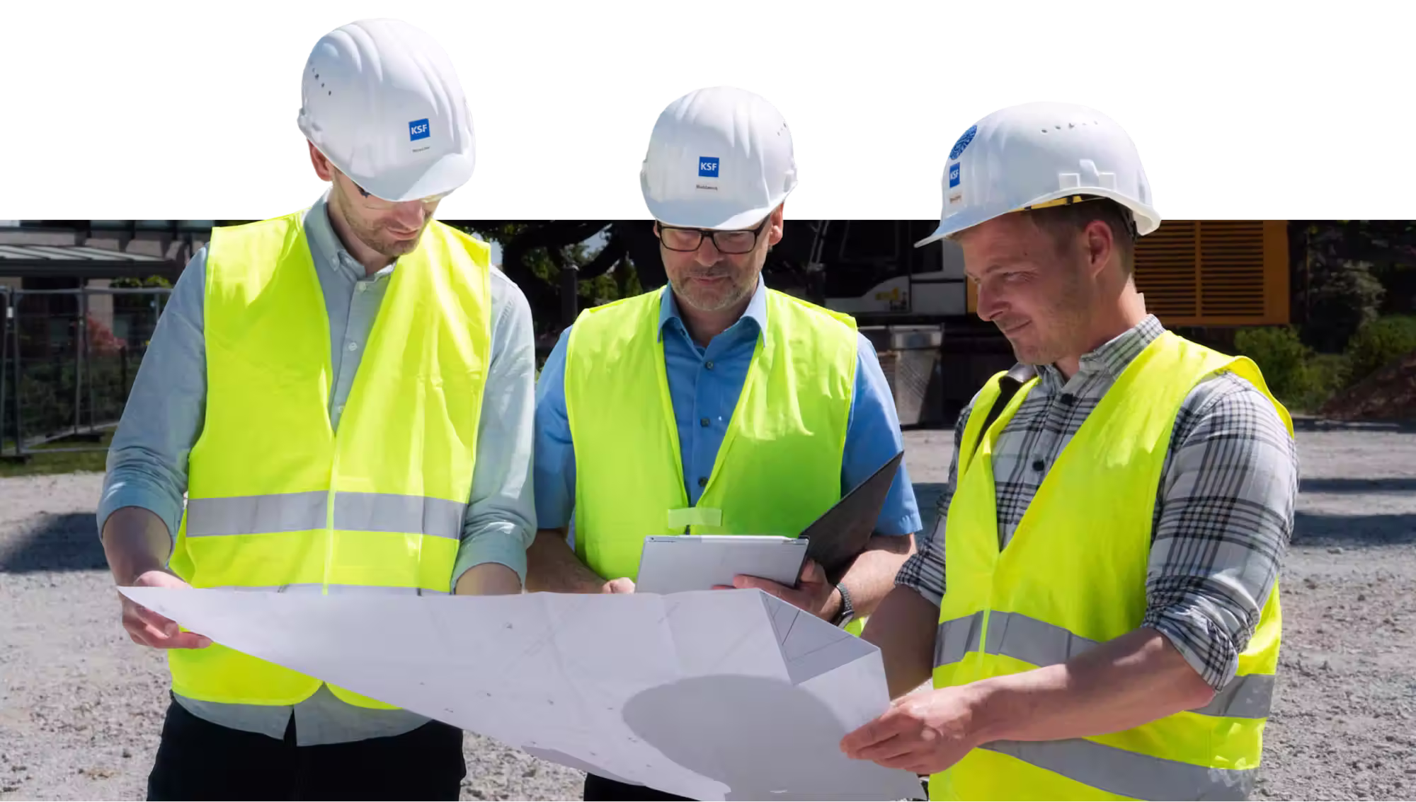 Drei Bauarbeiter mit weißen Helmen und gelben Warnwesten besprechen Pläne auf einer Baustelle.