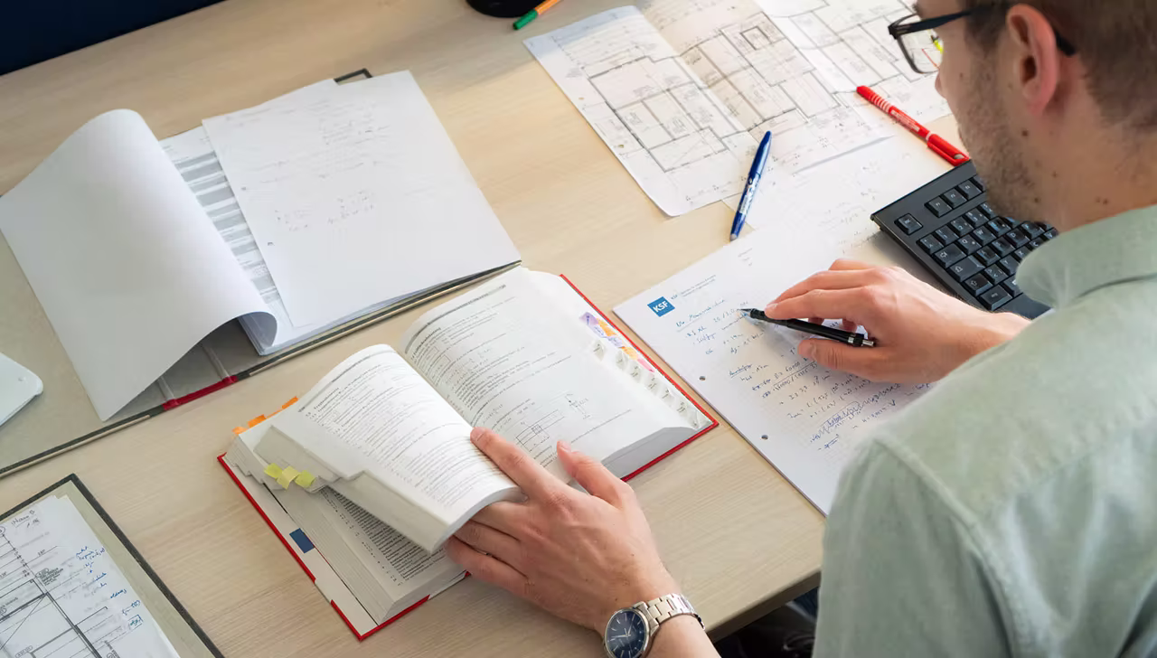 Person sitzt an einem Schreibtisch, liest ein Buch und macht sich Notizen auf Papier neben einer Tastatur und technischen Zeichnungen.