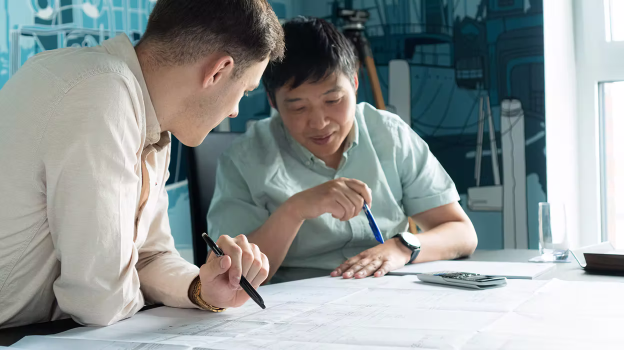 Zwei Männer diskutieren über Baupläne an einem Tisch, während sie Stifte halten.
