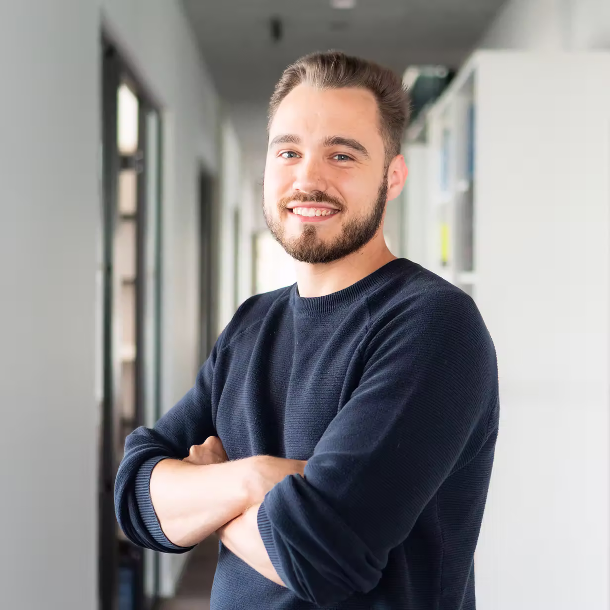 Junger Mann mit Bart und blauem Pullover steht mit verschränkten Armen in einem hellen Flur und lächelt.