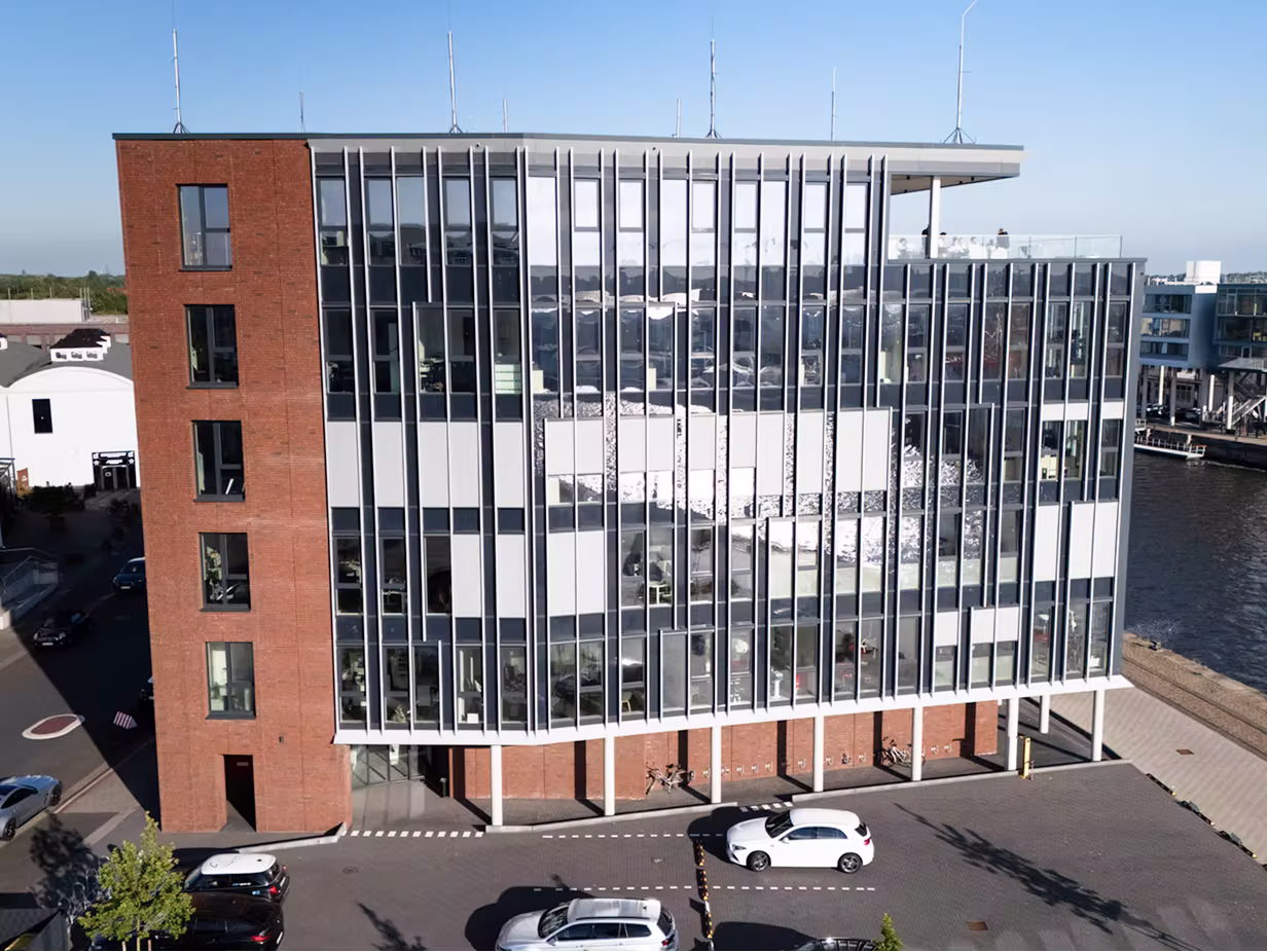 Modernes Bürogebäude mit Glasfassade und roten Backsteinwänden neben einer Wasserstraße, Autos auf dem Parkplatz davor.