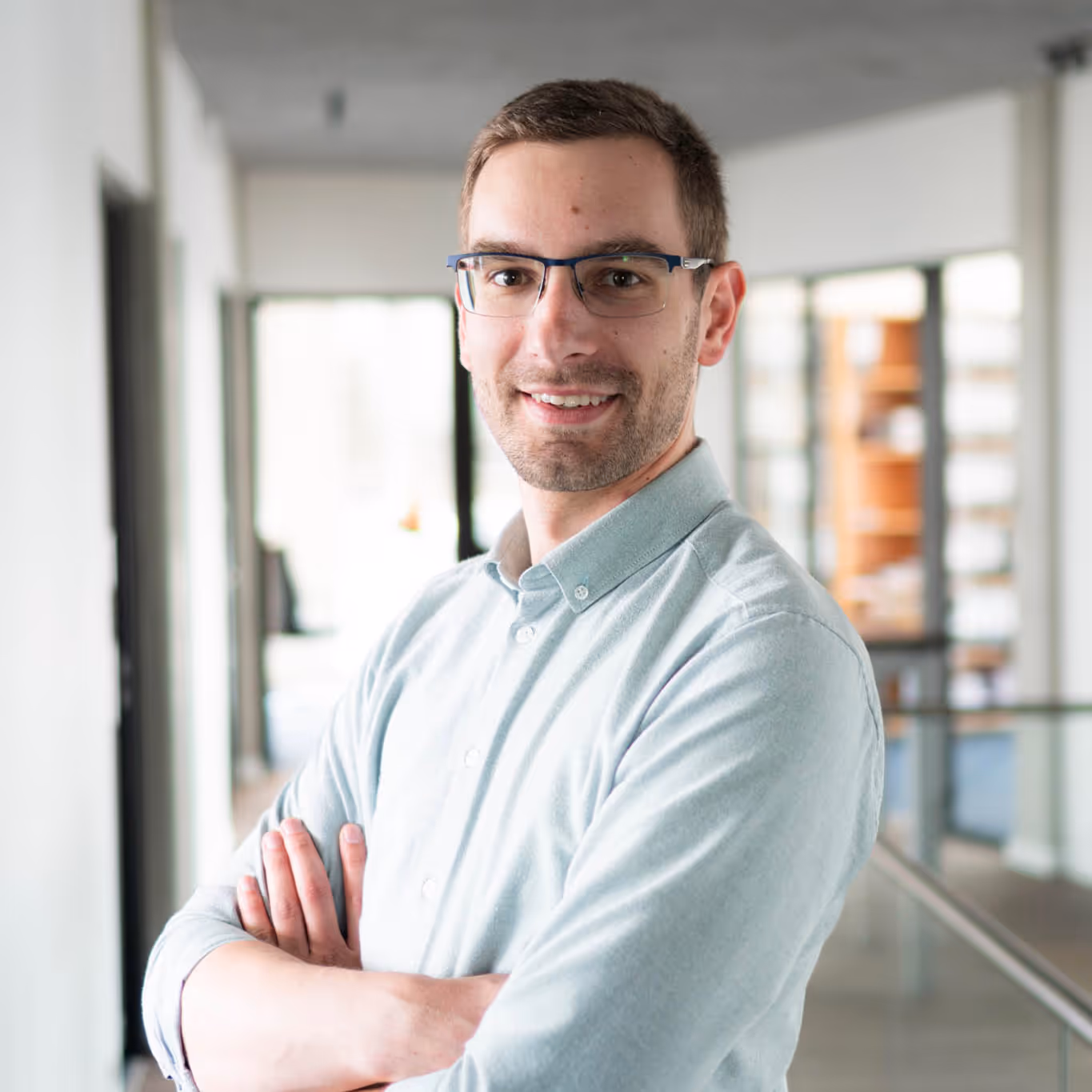 Ein lächelnder Mann mit Brille und hellblauem Hemd steht mit verschränkten Armen in einem modernen Büro.