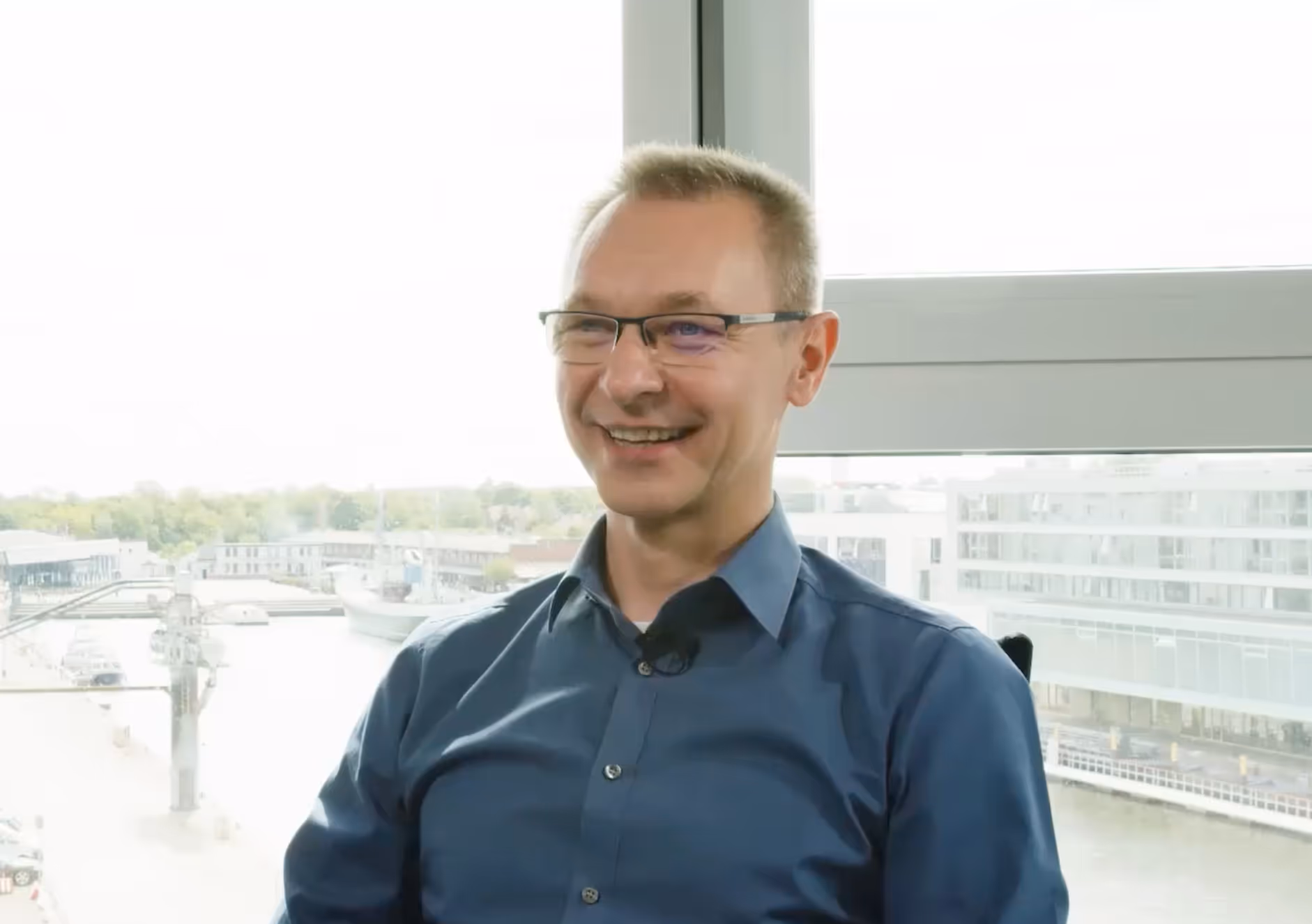 Lächelnder Mann mit Brille und blauem Hemd sitzt vor einem Fenster mit Blick auf einen Hafen.