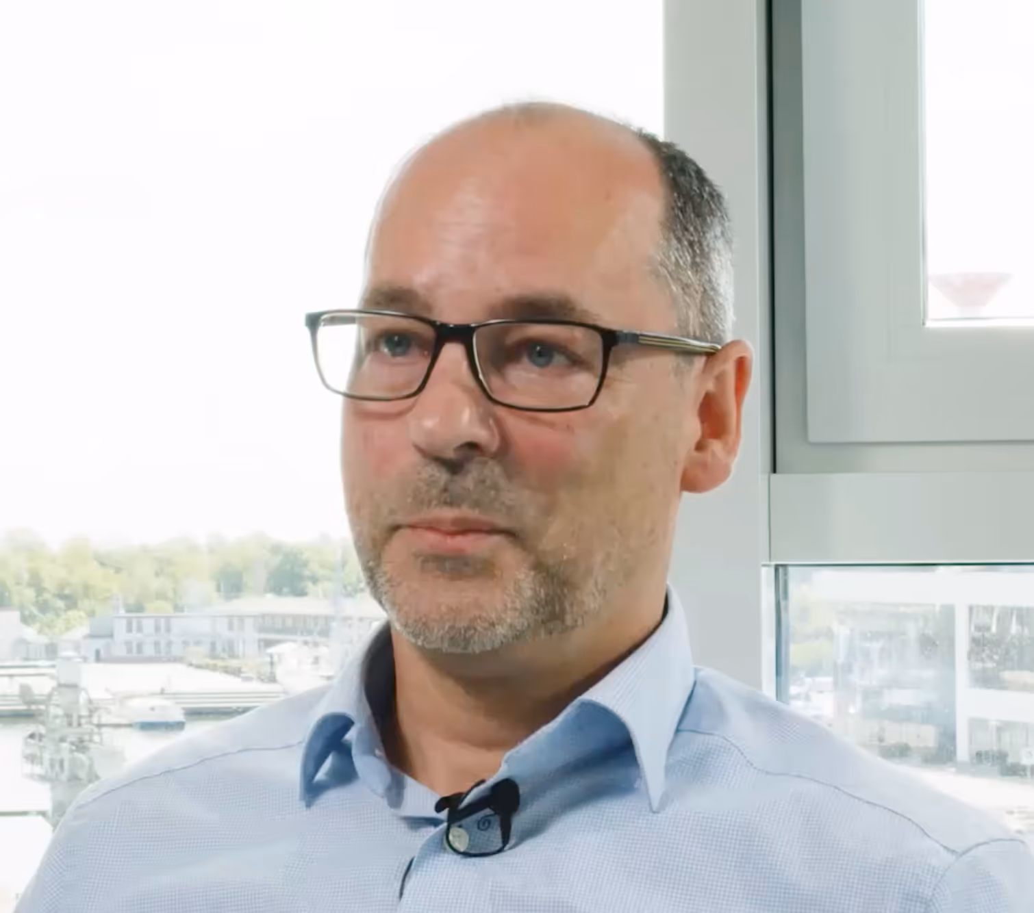 Mann mit Brille und Bart in einem hellen Raum vor einem Fenster mit Blick auf Gebäude und Bäume.