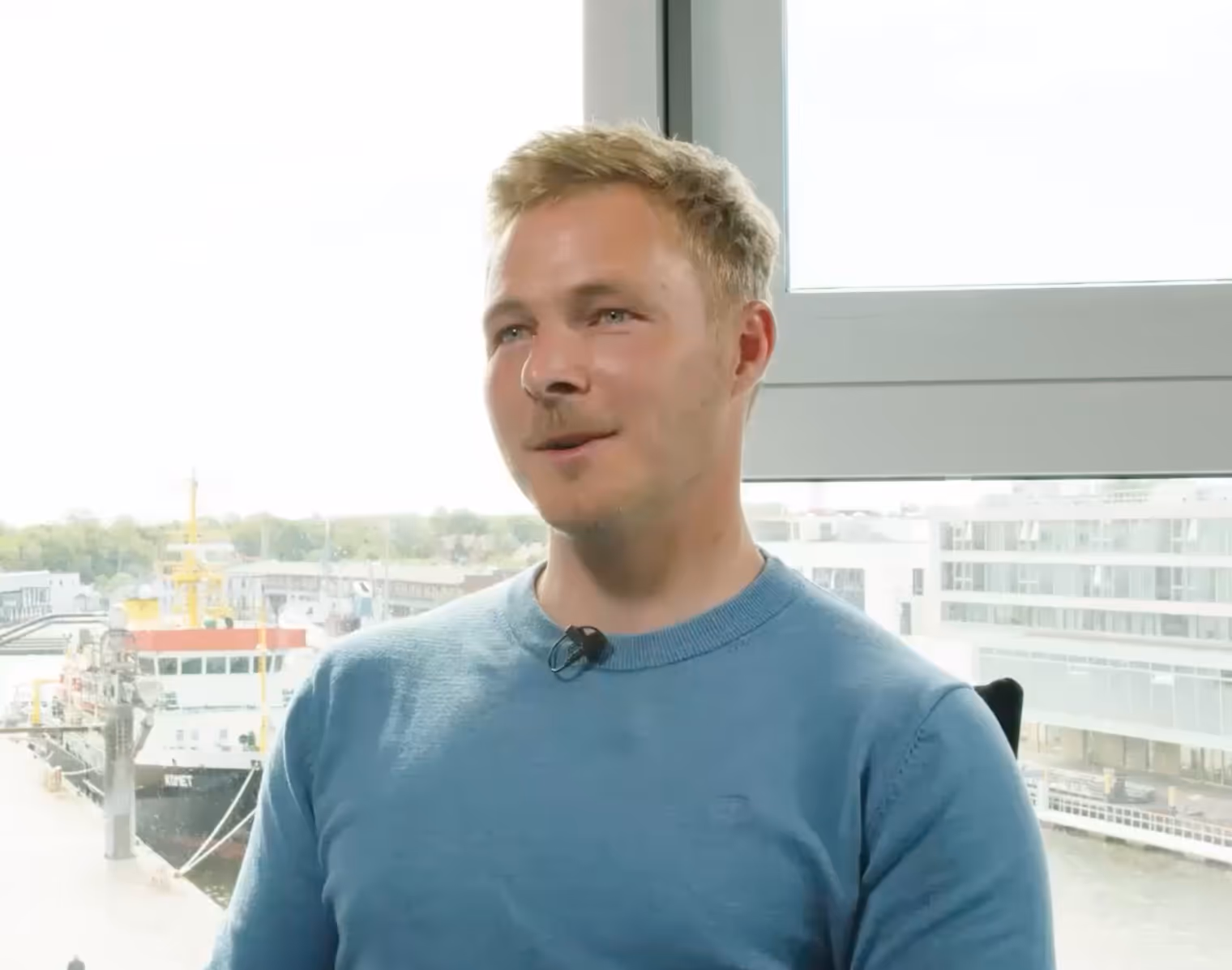 Junger Mann mit blondem Haar und blauem Pullover sitzt vor einem großen Fenster mit Blick auf Hafen und Schiffe.