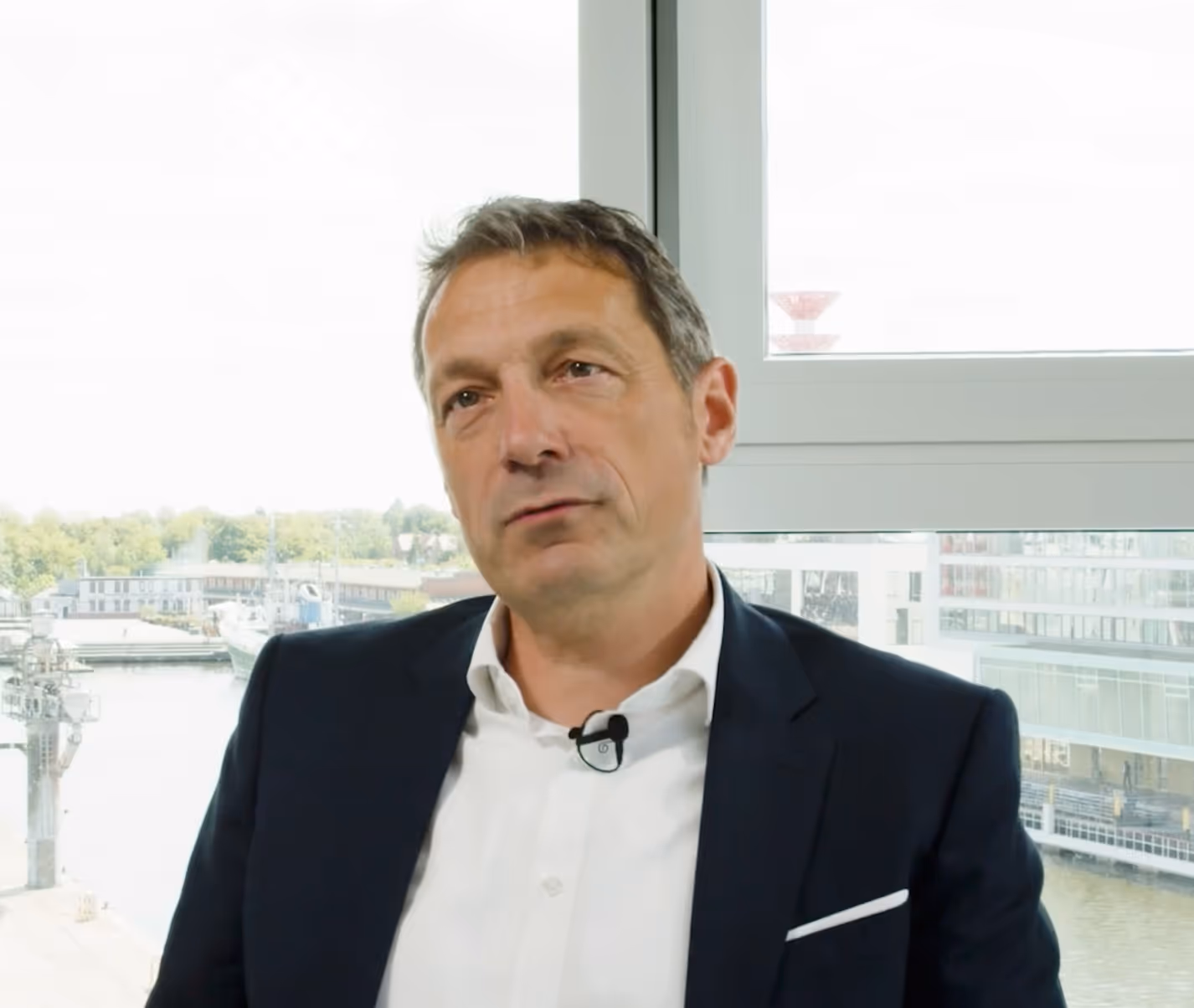 Ein Mann mit Anzug und weißem Hemd sitzt vor einem Fenster mit Blick auf einen Hafen.