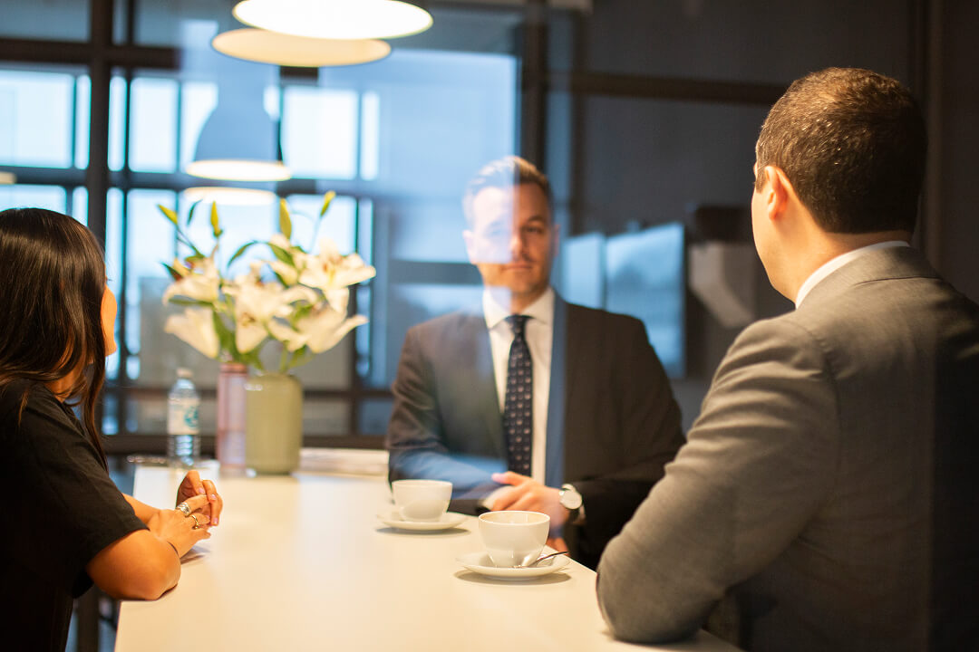 Drei Geschäftspersonen sitzen an einem Tisch in einem modernen Büro, zwei Männer in Anzügen und eine Frau mit langen dunklen Haaren.