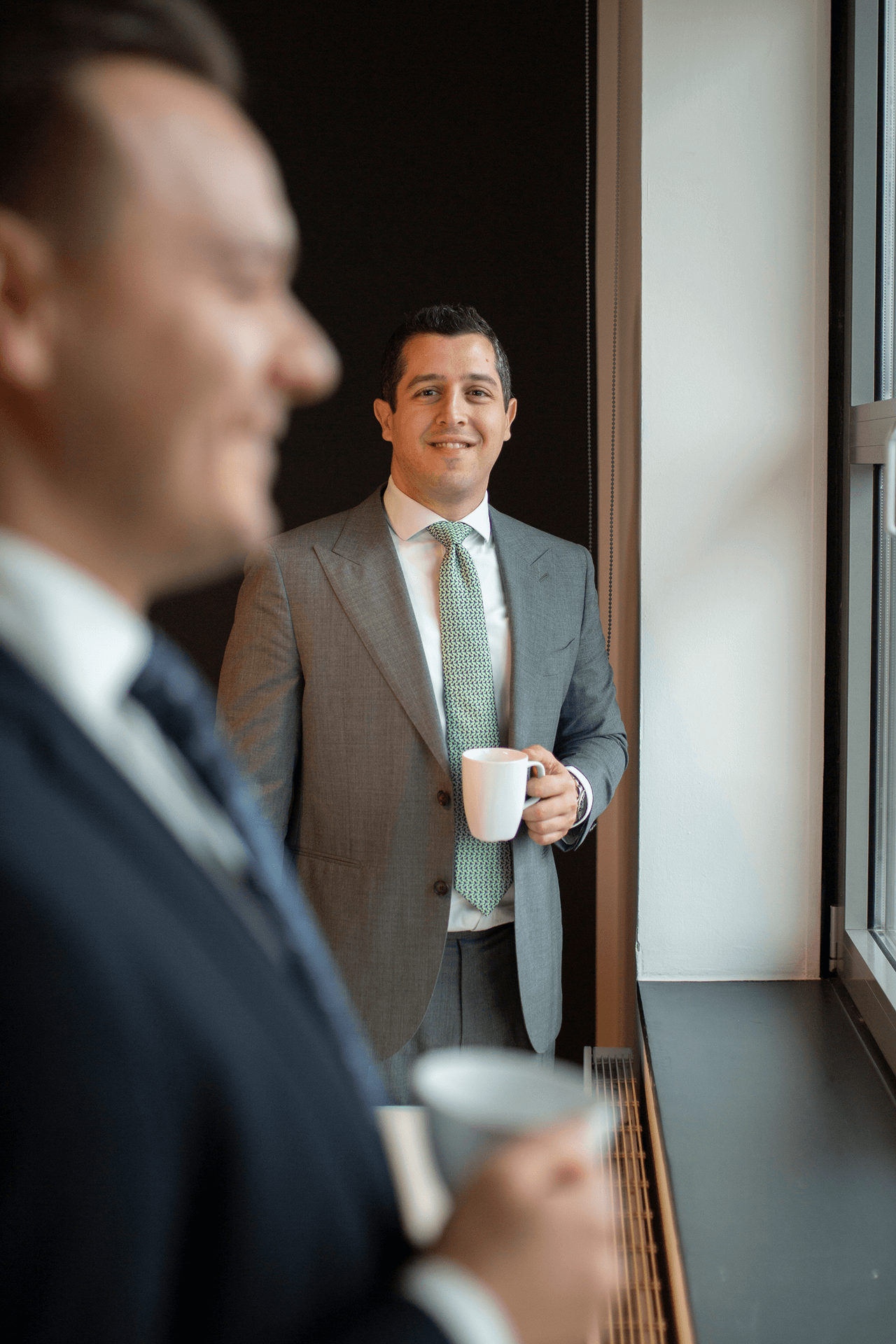 Zwei Männer in Anzügen stehen nebeneinander, einer im Vordergrund unscharf, der andere steht lächelnd am Fenster mit einer Tasse in der Hand.
