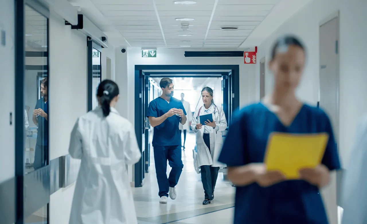 A busy hospital corridor with diverse medical professionals, including doctors in white coats and nurses in blue scrubs, walking and discussing patient care.