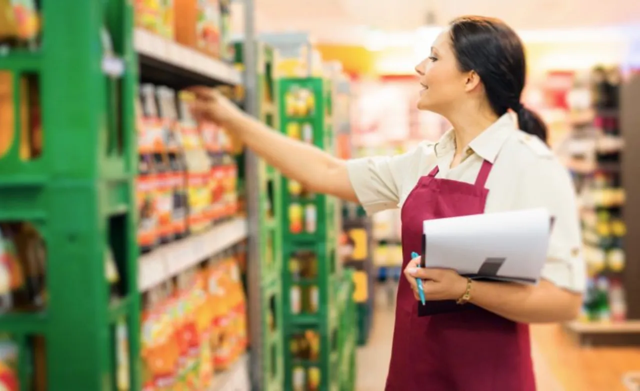A female retail employee in a red apron holding a clipboard while checking inventory and organizing products on shelves in a grocery store.