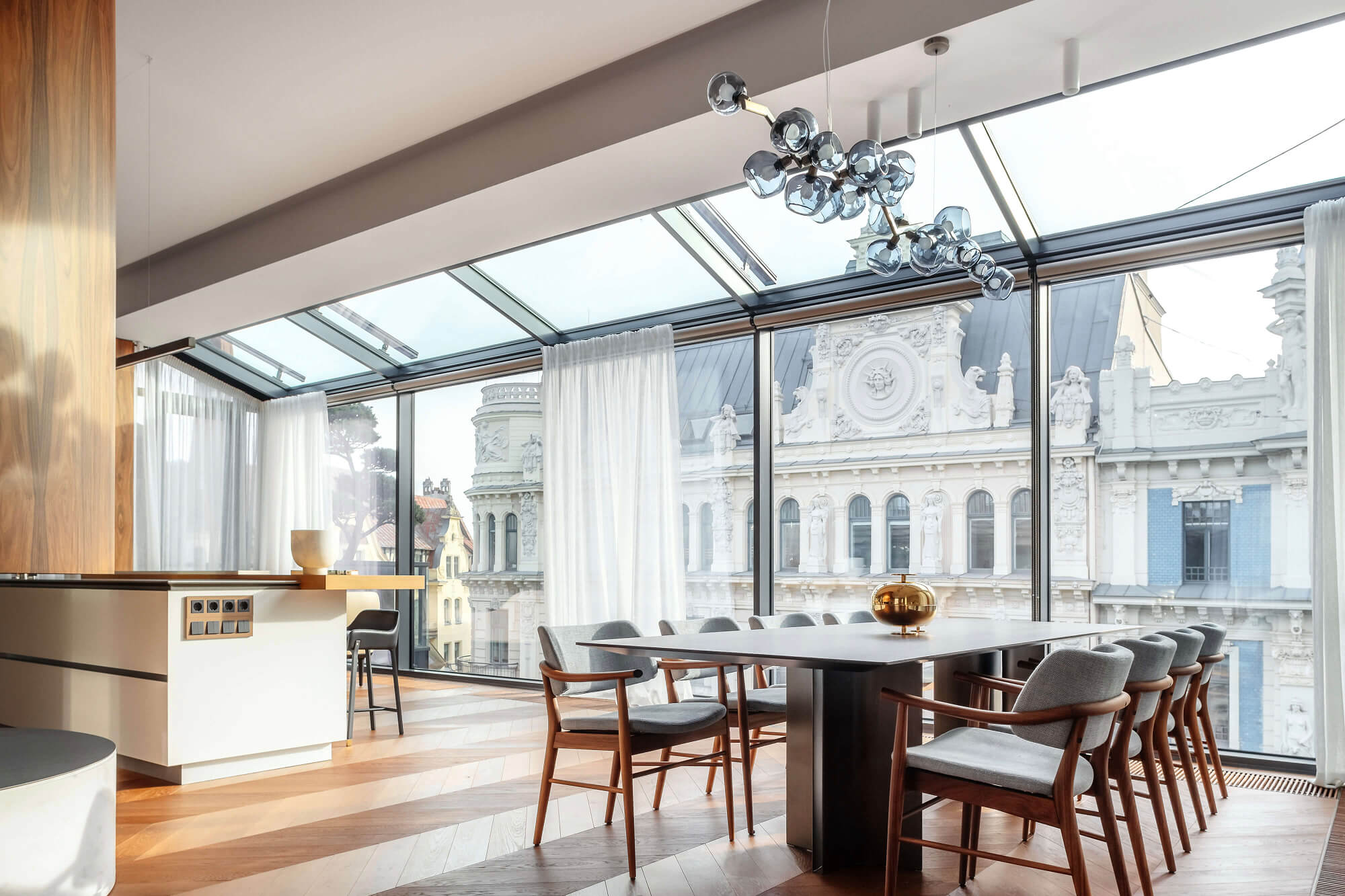 Modern dining area with wooden floor, large table surrounded by gray cushioned chairs, glass ceiling and wall overlooking ornate historic building.