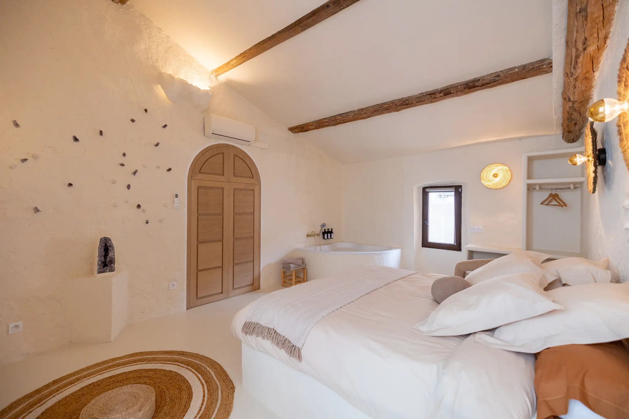 Chambre raffinée à La Garde, avec lit double aux draps crème, baignoire d’angle, murs texturés ornés de cristaux, poutres apparentes et éléments naturels comme un tapis en fibres tressées et une géode d’améthyste.