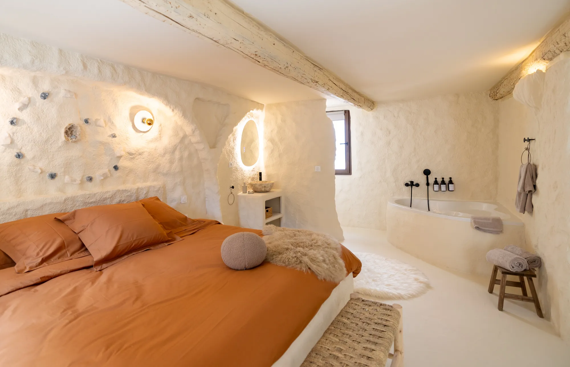 Chambre lumineuse à l’ambiance naturelle, avec un lit aux draps couleur terracotta, une baignoire d’angle intégrée dans un mur texturé, des serviettes pliées, un miroir rétroéclairé et des poutres apparentes au plafond.