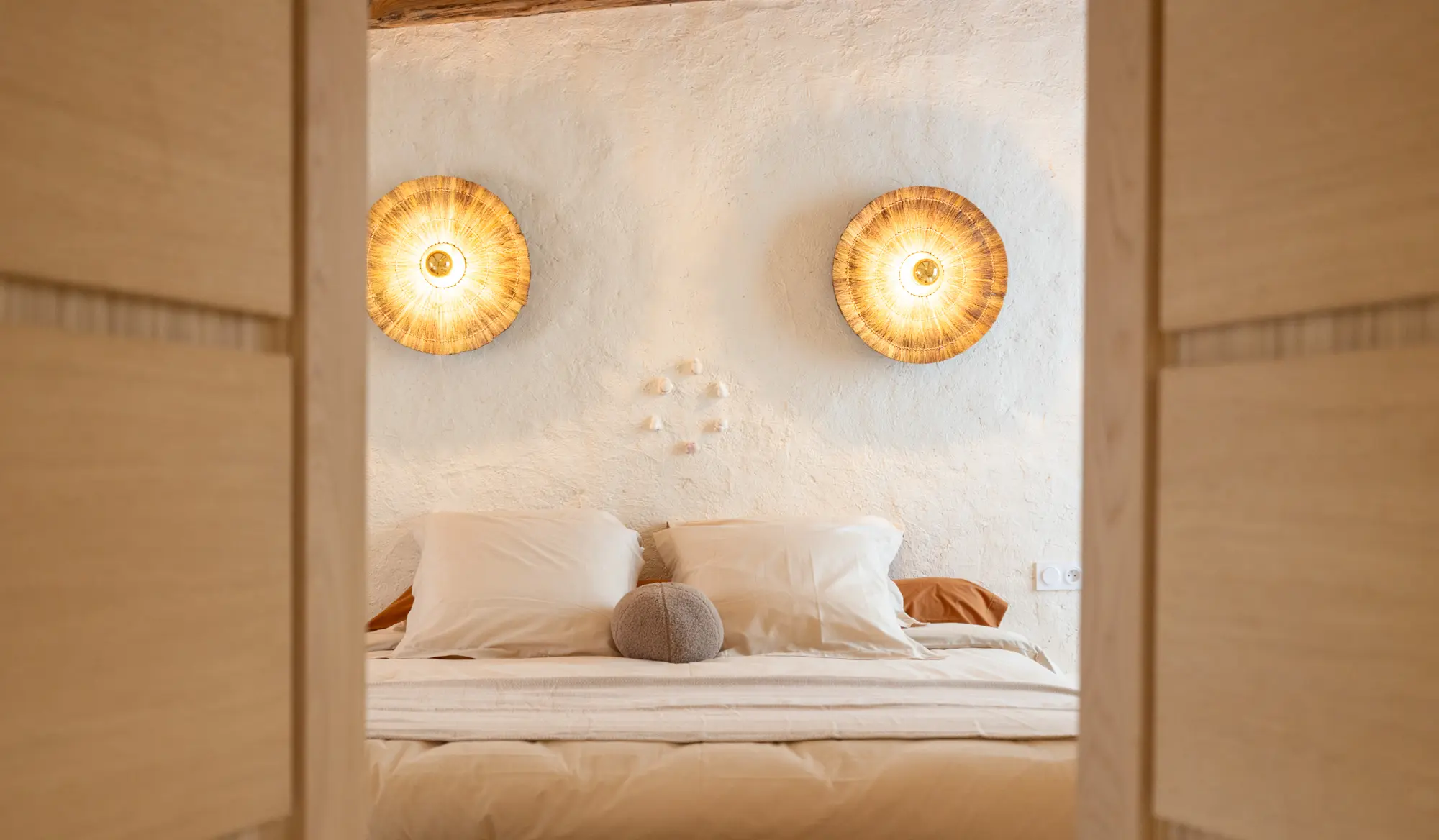 Vue à travers une porte entrouverte sur une chambre paisible de Casa Anka, située à La Garde, avec un lit aux tons neutres, deux appliques murales en fibres naturelles et une décoration murale minimaliste en pierres.