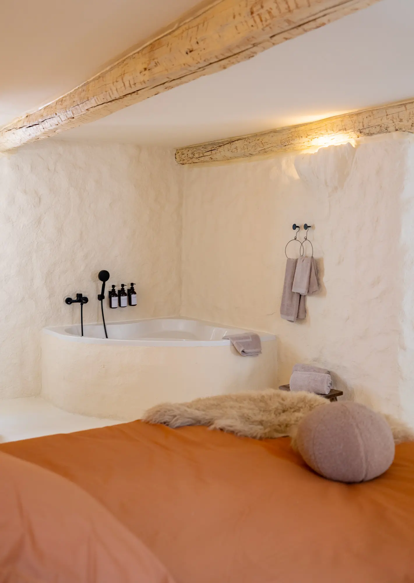 Espace cocooning à Casa Ananta, avec une baignoire d’angle blanche intégrée dans un mur texturé, des serviettes soigneusement pliées et des poutres en bois apparentes, créant une ambiance chaleureuse et naturelle.