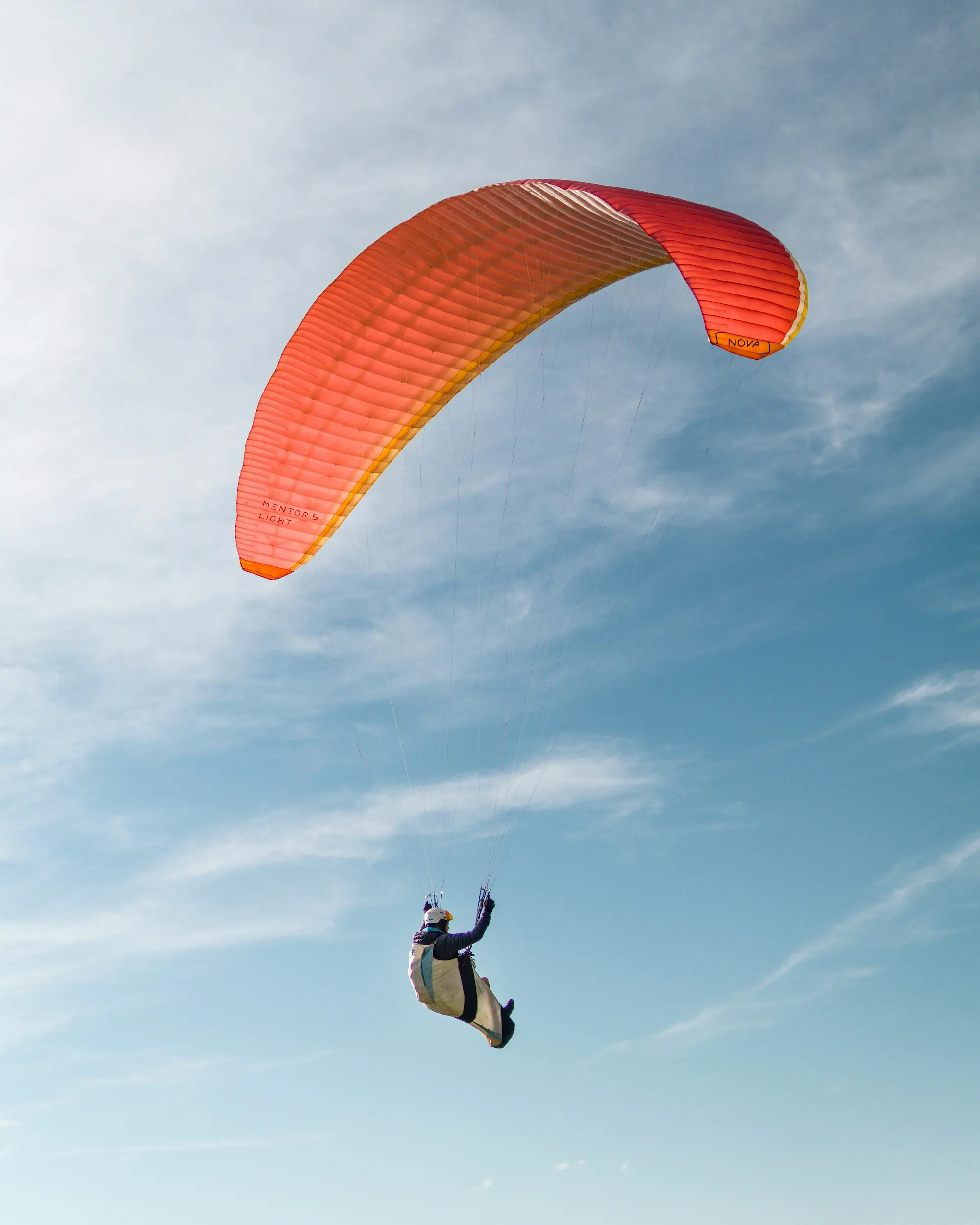 Parapentiste en plein vol sous une voile rouge-orange, évoluant dans un ciel bleu parsemé de fins nuages.