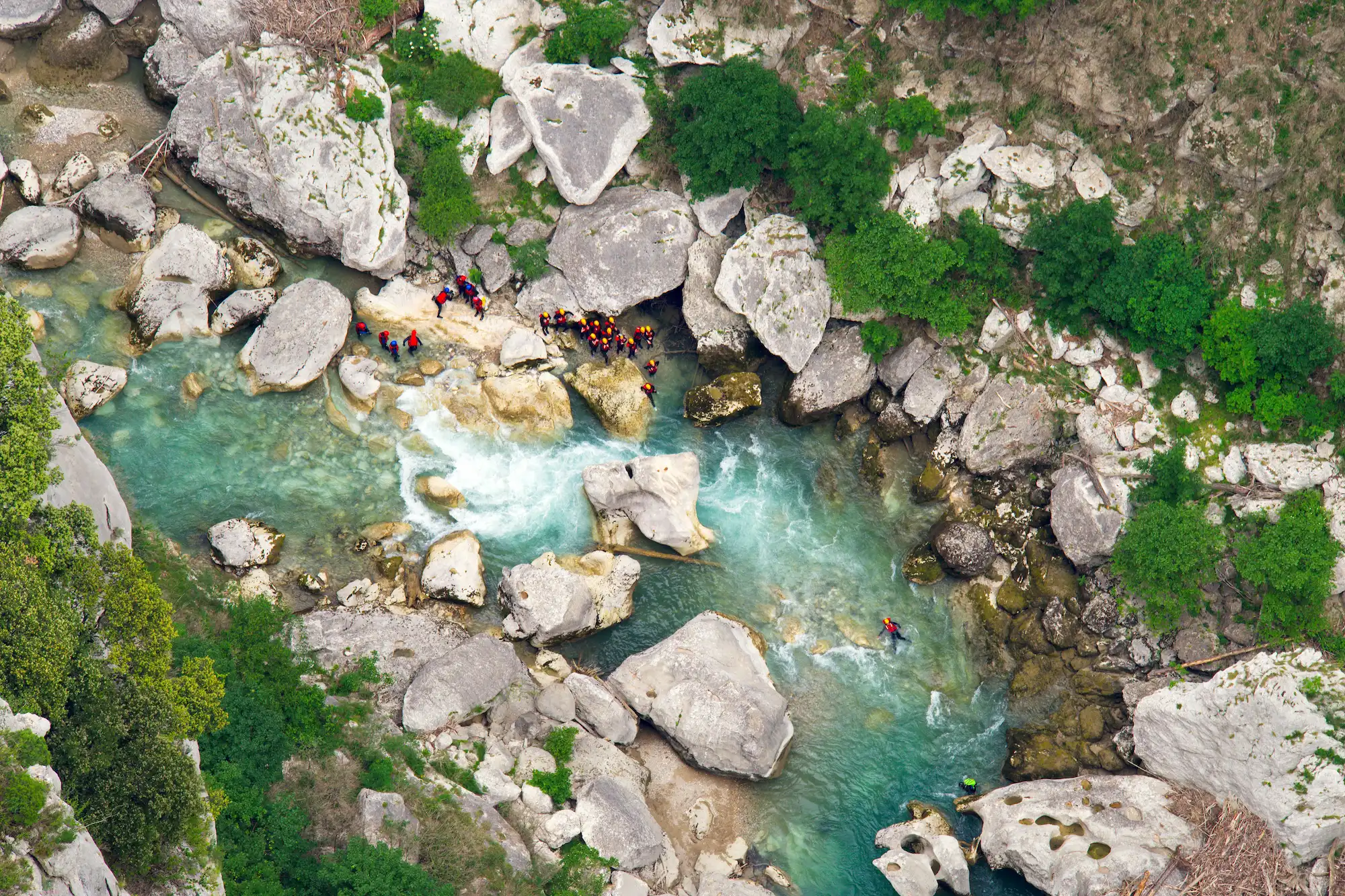 Vue aérienne d’un groupe de personnes en combinaison de canyoning explorant un cours d’eau turquoise bordé de gros rochers et de végétation dense dans les gorges du Verdon.