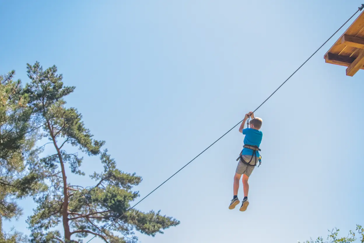 Un garçon en tenue de sport glisse le long d'une tyrolienne en plein ciel bleu, attaché par un harnais de sécurité, avec des arbres en arrière-plan.