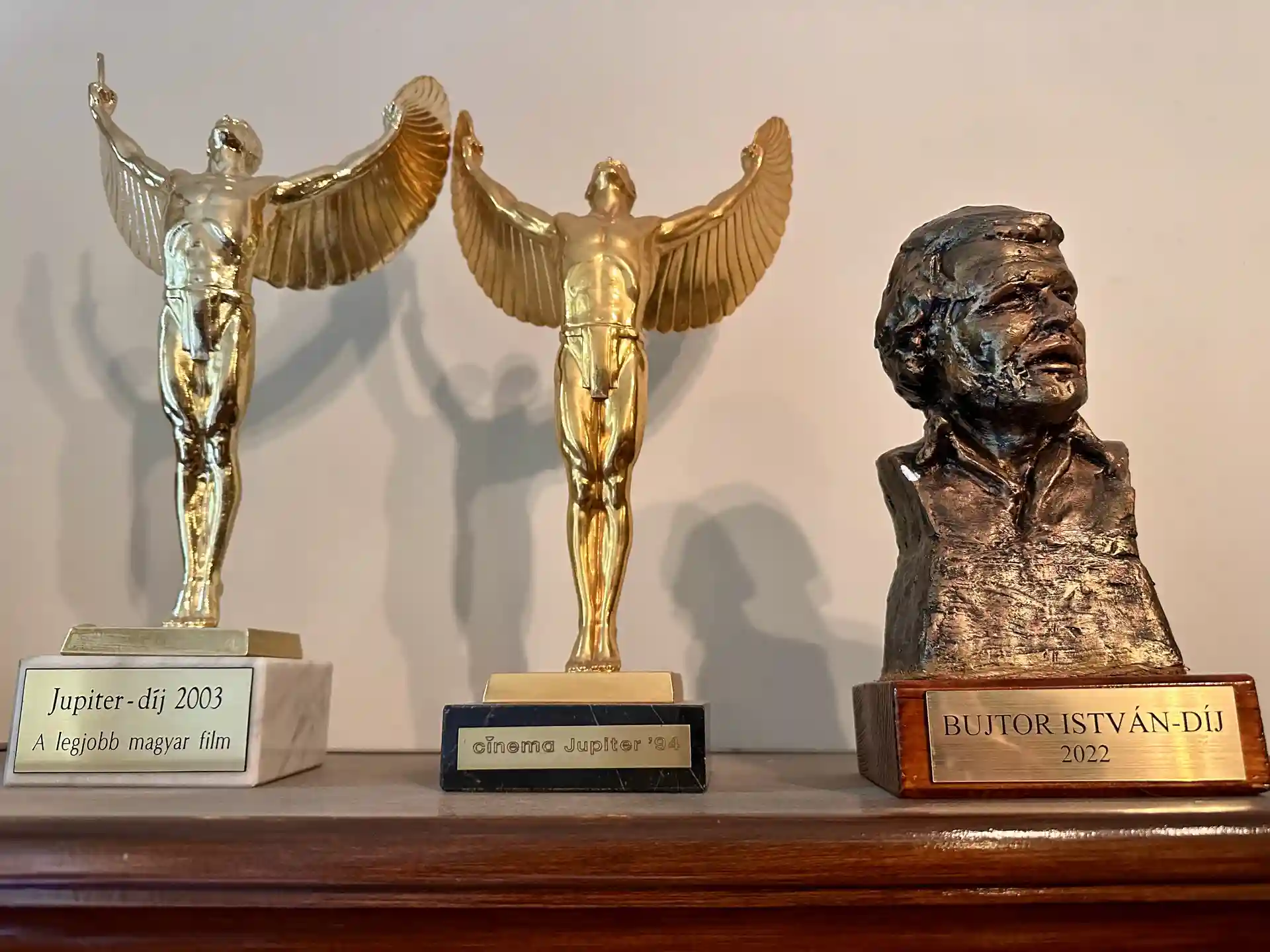Three awards on a wooden shelf: two golden winged male figures with plaques reading Jupiter Award 2003 and Cinema Jupiter '94, and a bronze bust with a plaque reading Bujtor István Award 2022.