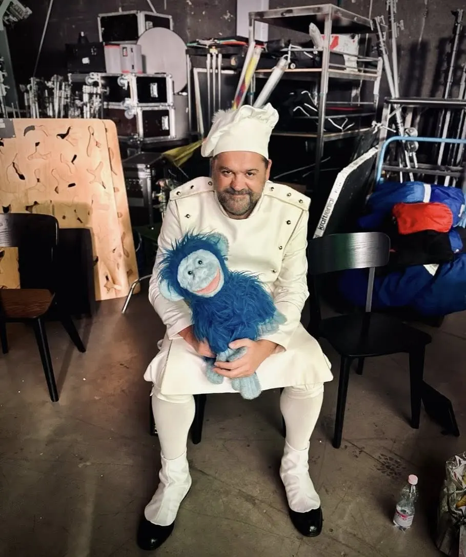 Man dressed as a chef sitting on a black chair holding a blue furry monkey puppet, surrounded by equipment cases and chairs.