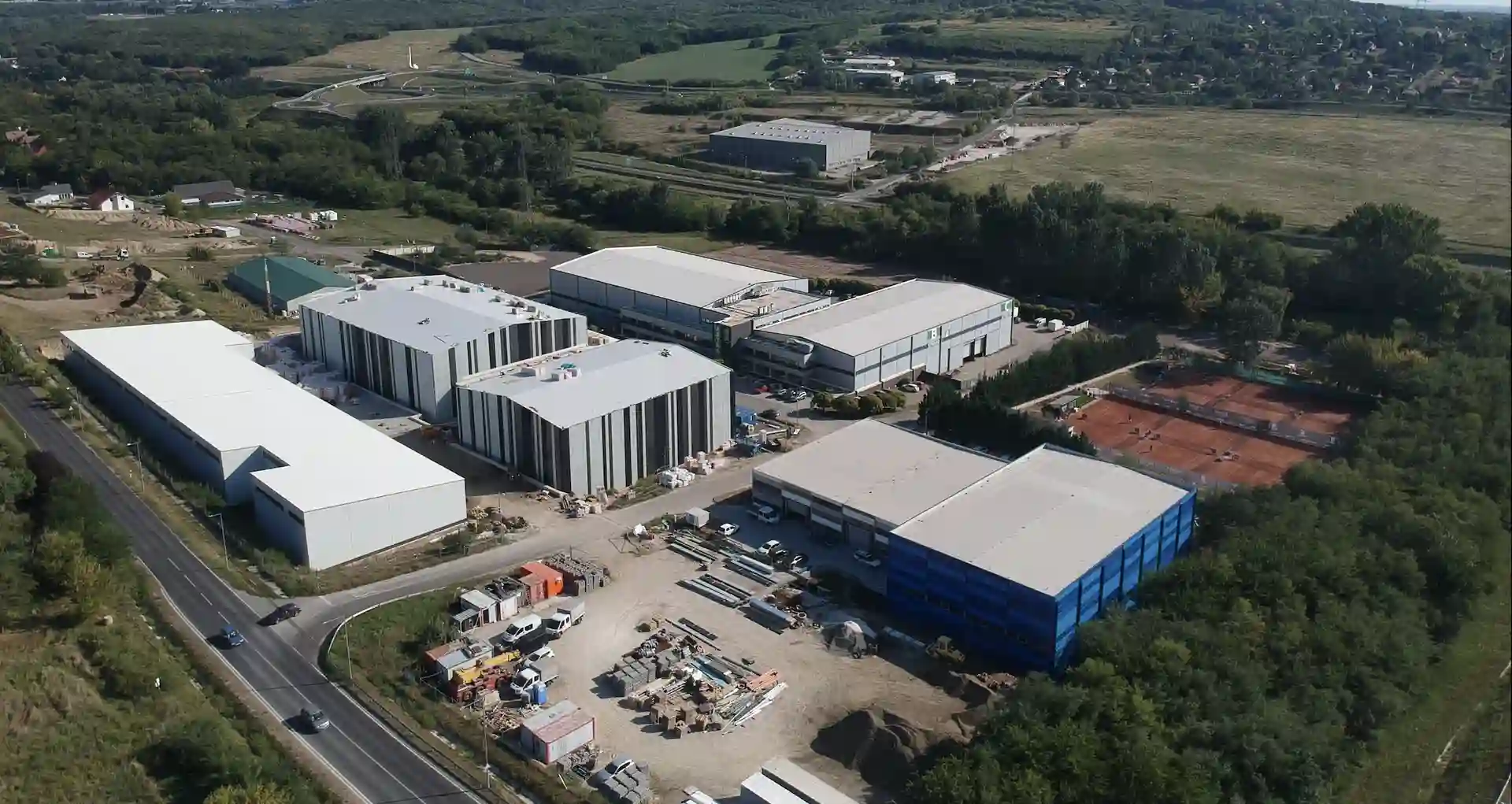 Aerial view of an industrial complex with multiple large warehouse buildings, surrounded by trees and a road with vehicles.