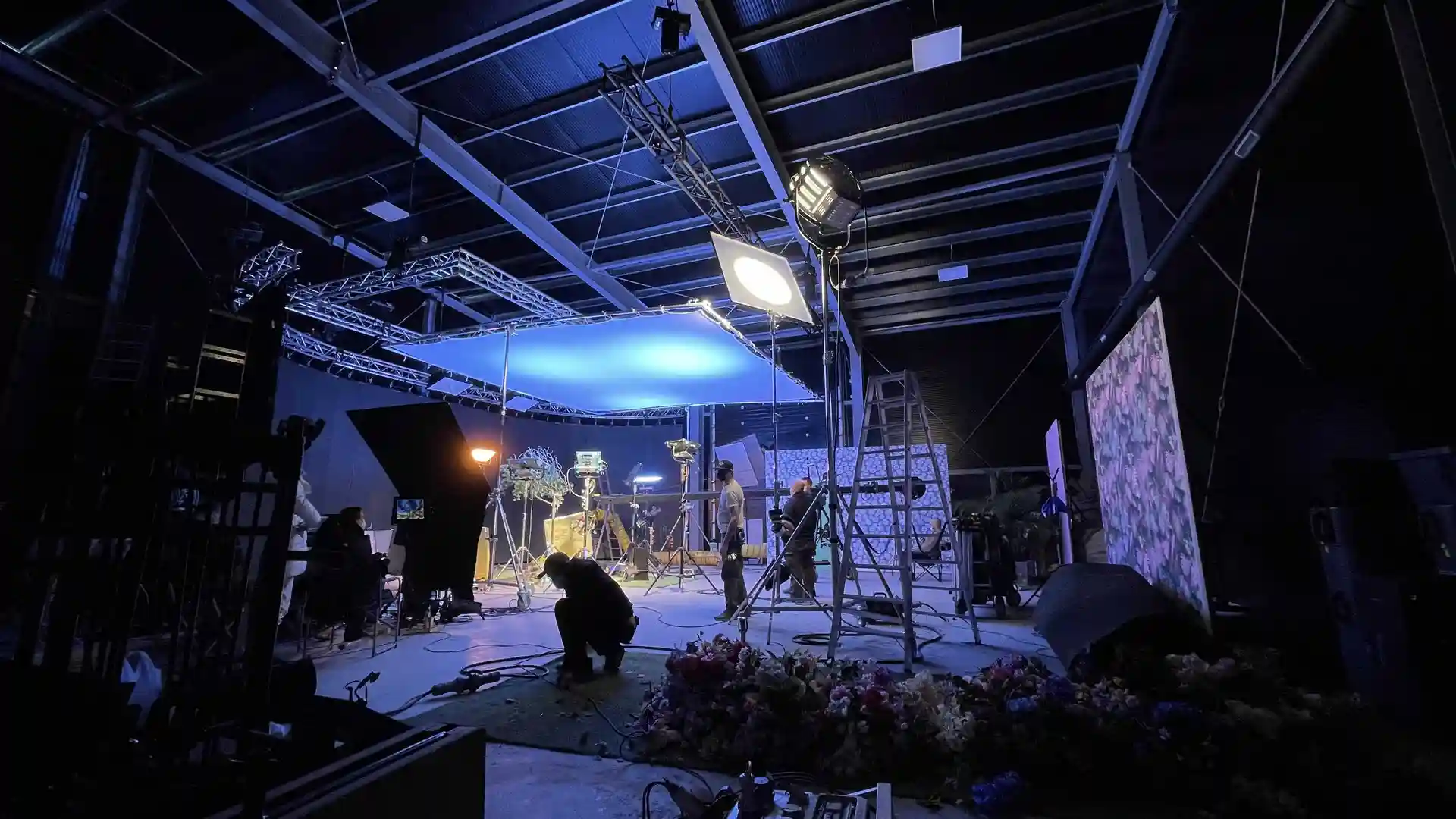 Indoor film or photo studio setup with bright lights, equipment stands, and crew members working under a blue-lit ceiling panel.