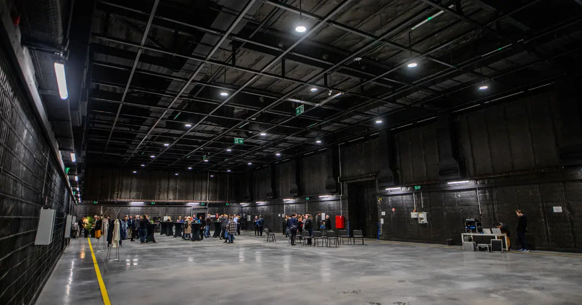 Large industrial sound stage with black walls and ceiling, people gathered around high tables, and chairs near the center.