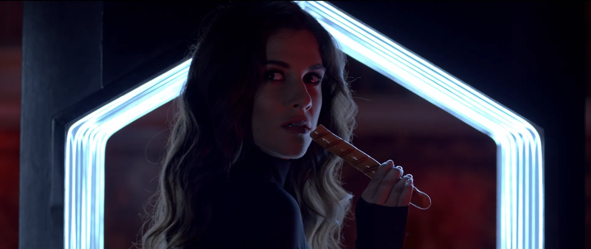 Woman with long wavy hair holding a grilled sausage near her mouth against a dark background with glowing geometric light frame.