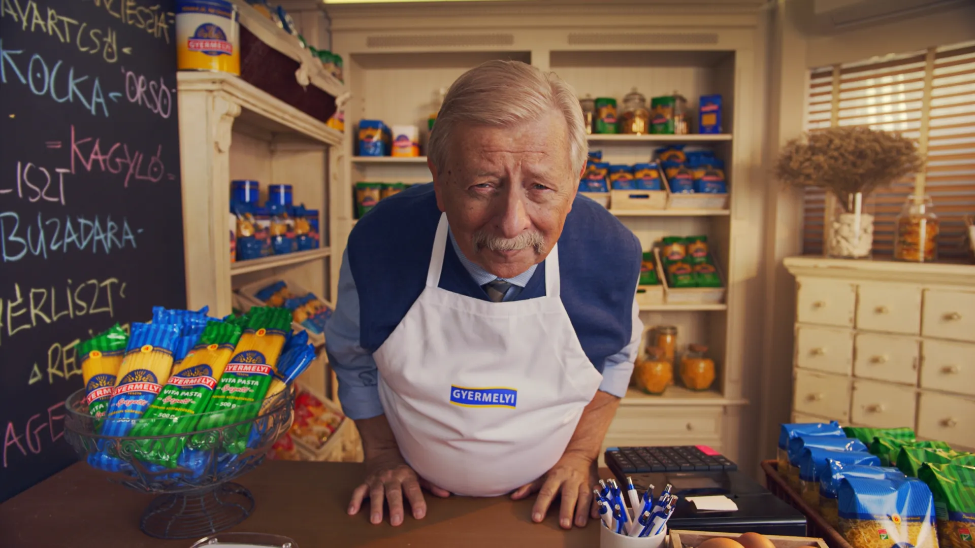 Elderly man wearing a white apron that says GYERMELYI leaning forward behind a counter with pasta packages in a shop.