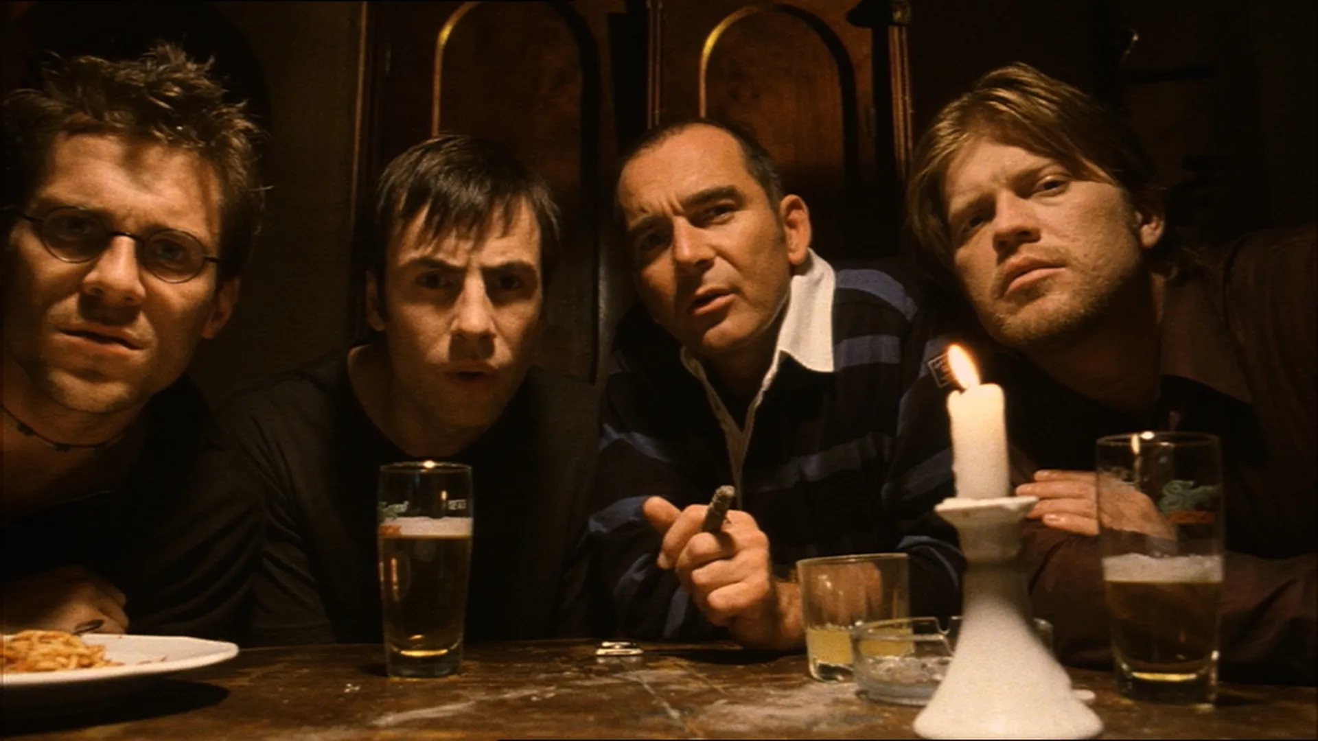 Four men sitting closely at a dimly lit table with drinks, a candle, and a plate of food.