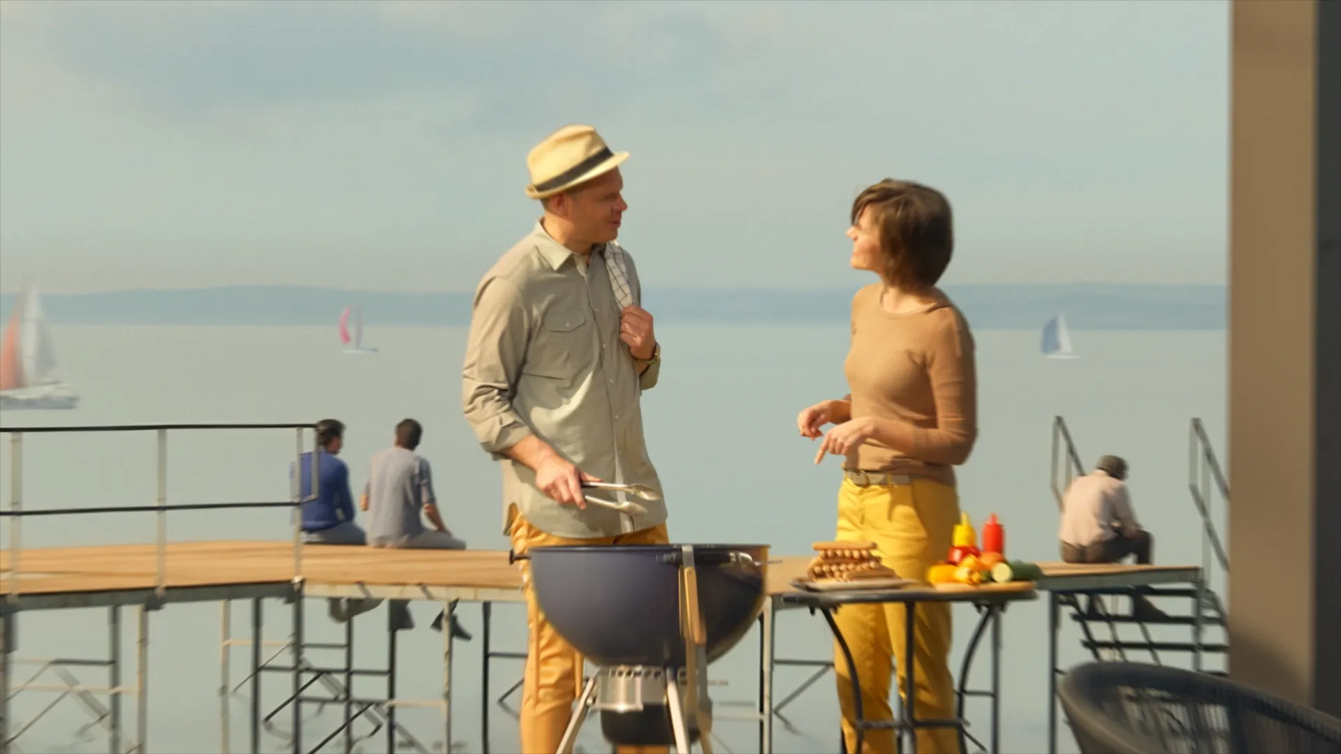 A man and a woman stand by a barbecue grill near a lake pier with sailboats in the background, preparing food on a sunny day.